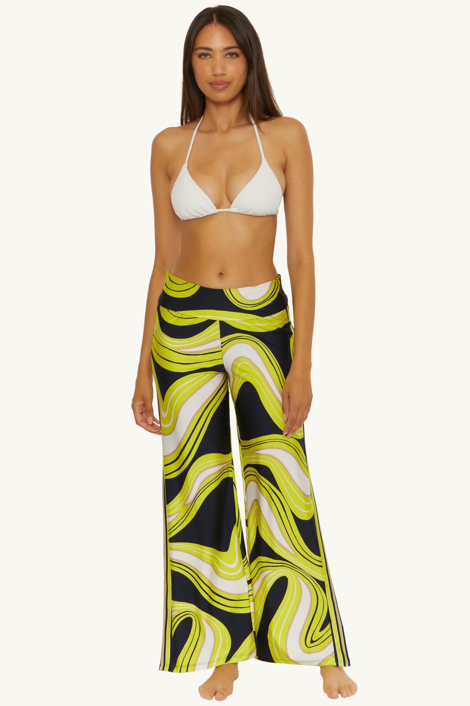 A woman stands barefoot against a plain background, wearing a white bikini top and the Serena Swirl Swim Cover-Up Pant, featuring a high-waisted, wide-leg design with bold yellow, black, and white swirls.