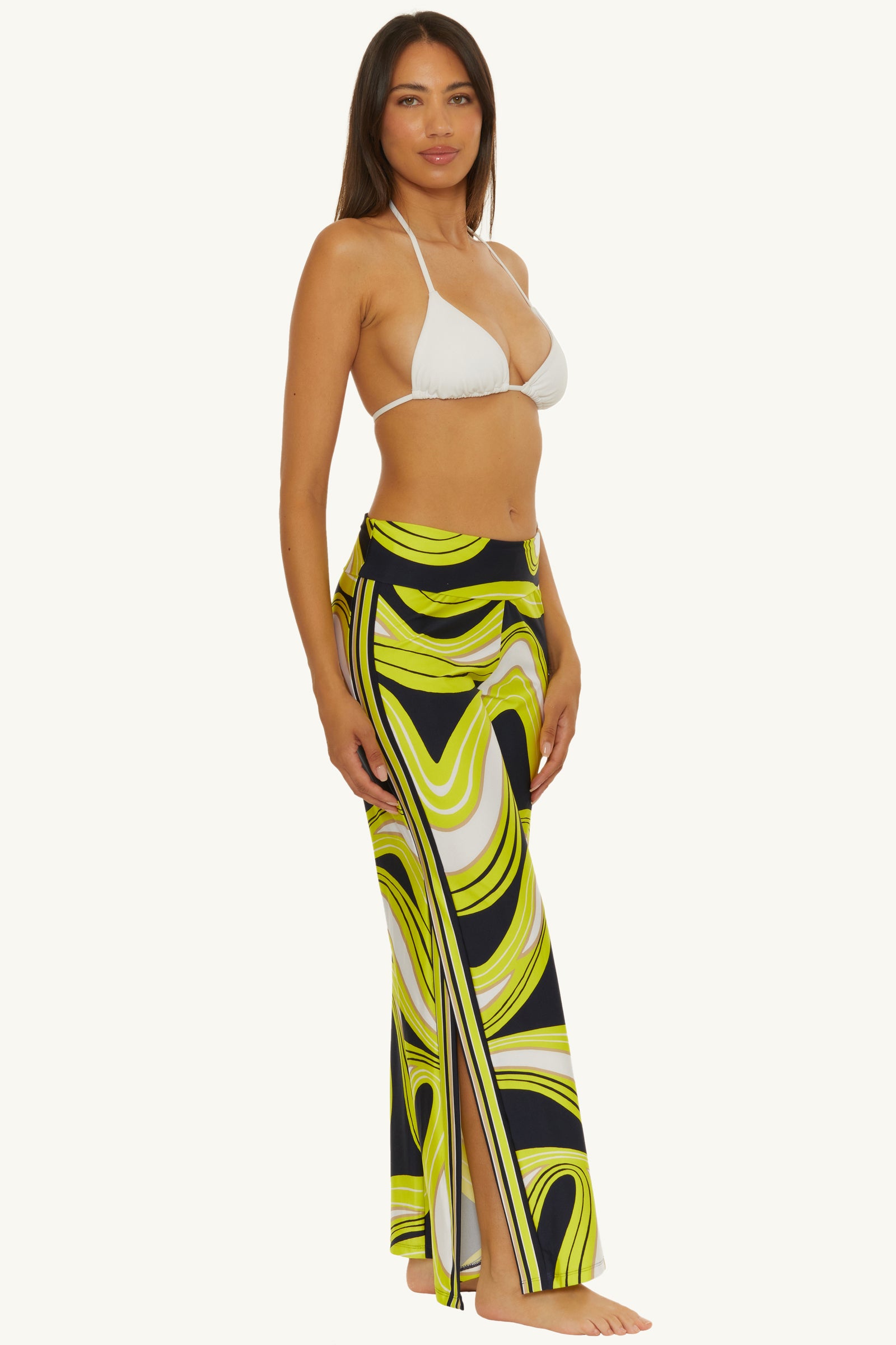 A woman stands barefoot wearing a white bikini top and the Swim Cover-Up Pant - Serena Swirl, featuring high-waisted, wide-leg pants with a bold yellow, black, and white swirl pattern and a side slit. She faces slightly to the side, looking at the camera.