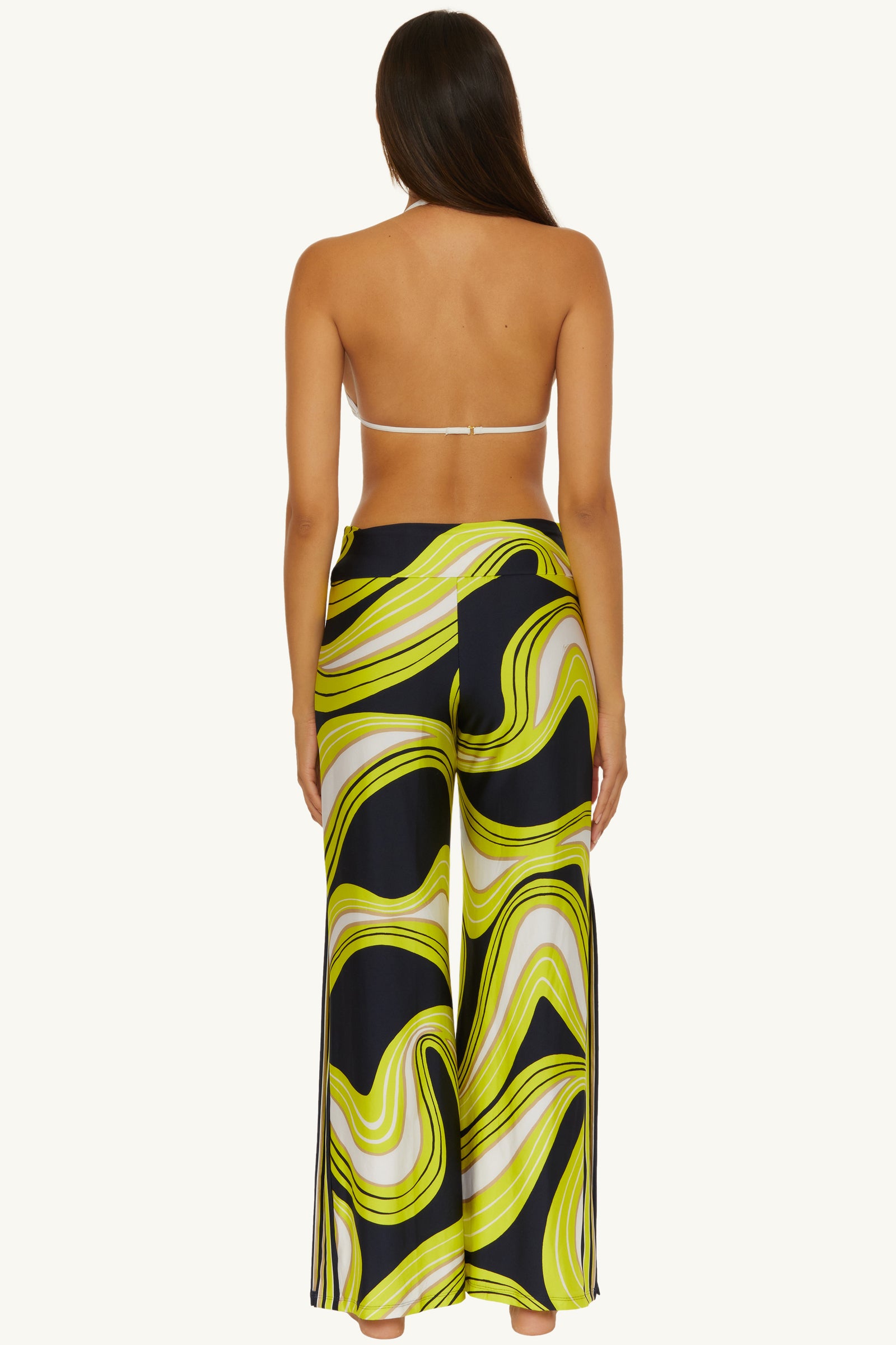 Woman stands barefoot with her back to the camera, wearing a white strapless bikini top and the Swim Cover-Up Pant - Serena Swirl, featuring a bold yellow, white, and black swirled pattern with a high-waisted, wide-leg design.