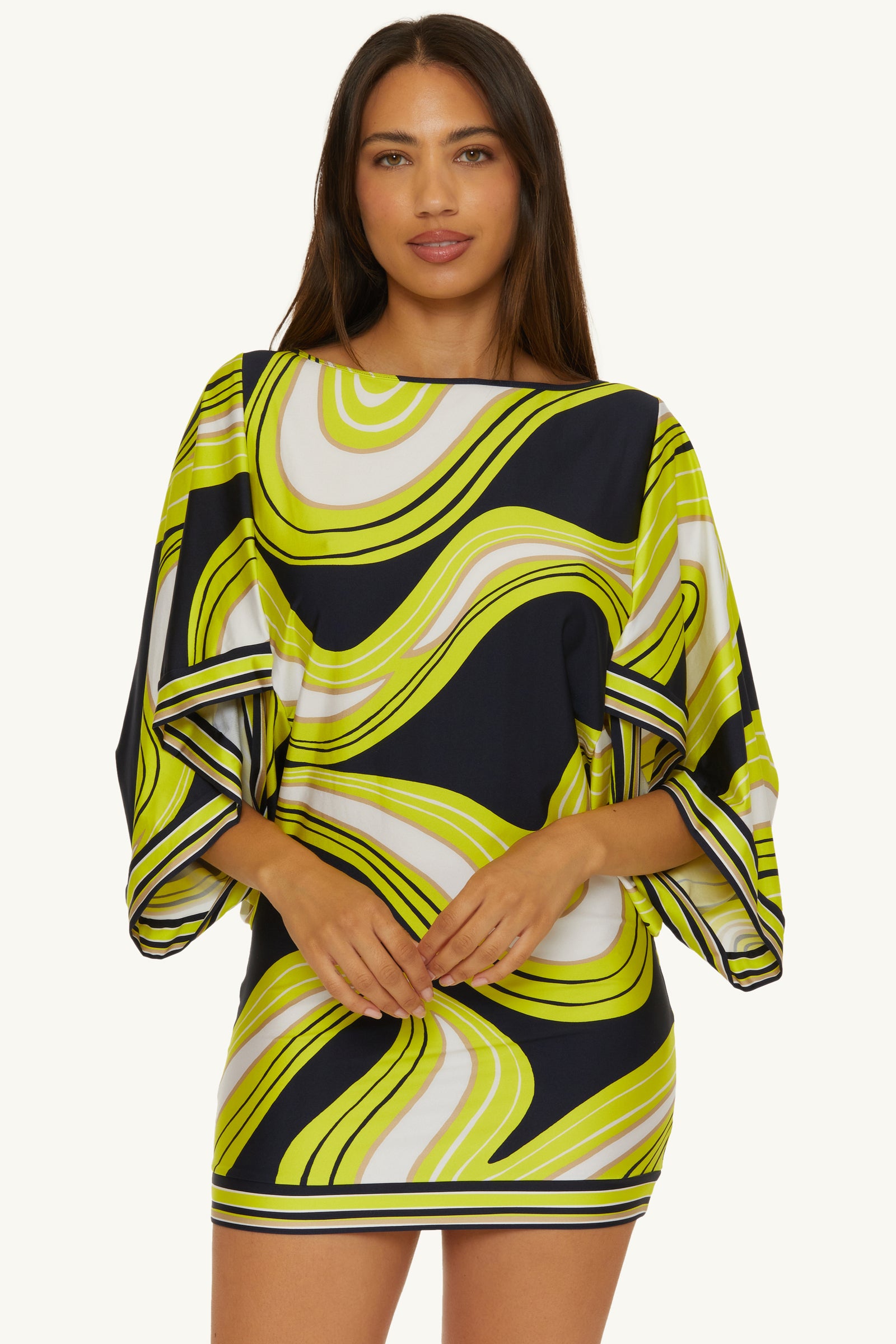 A woman stands facing forward with her hands clasped, wearing the Casablanca Tunic Cover-Up in Serena Swirl Swim, featuring a colorful, bold pattern and wide sleeves.