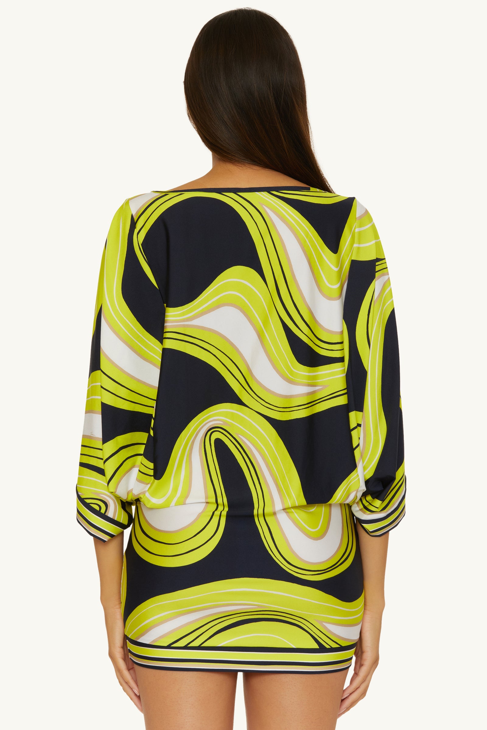 A woman with long brown hair faces away, wearing the Casablanca Tunic Cover-Up in Serena Swirl—a black, white, and yellow-green pattern with loose three-quarter sleeves and a fitted skirt.