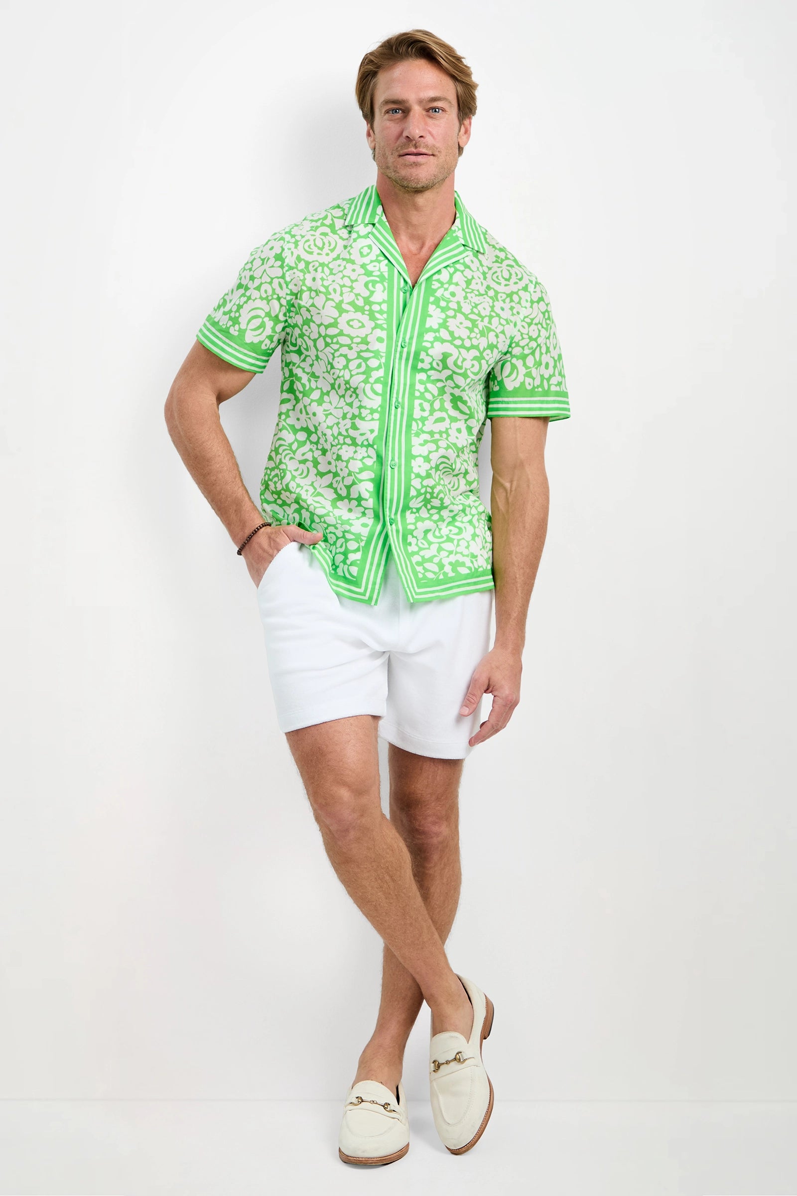A man poses against a white background wearing the Josue Short-Sleeve Shirt in Poolside Gossip, paired with white shorts and cream loafers, one leg crossed and arms relaxed.