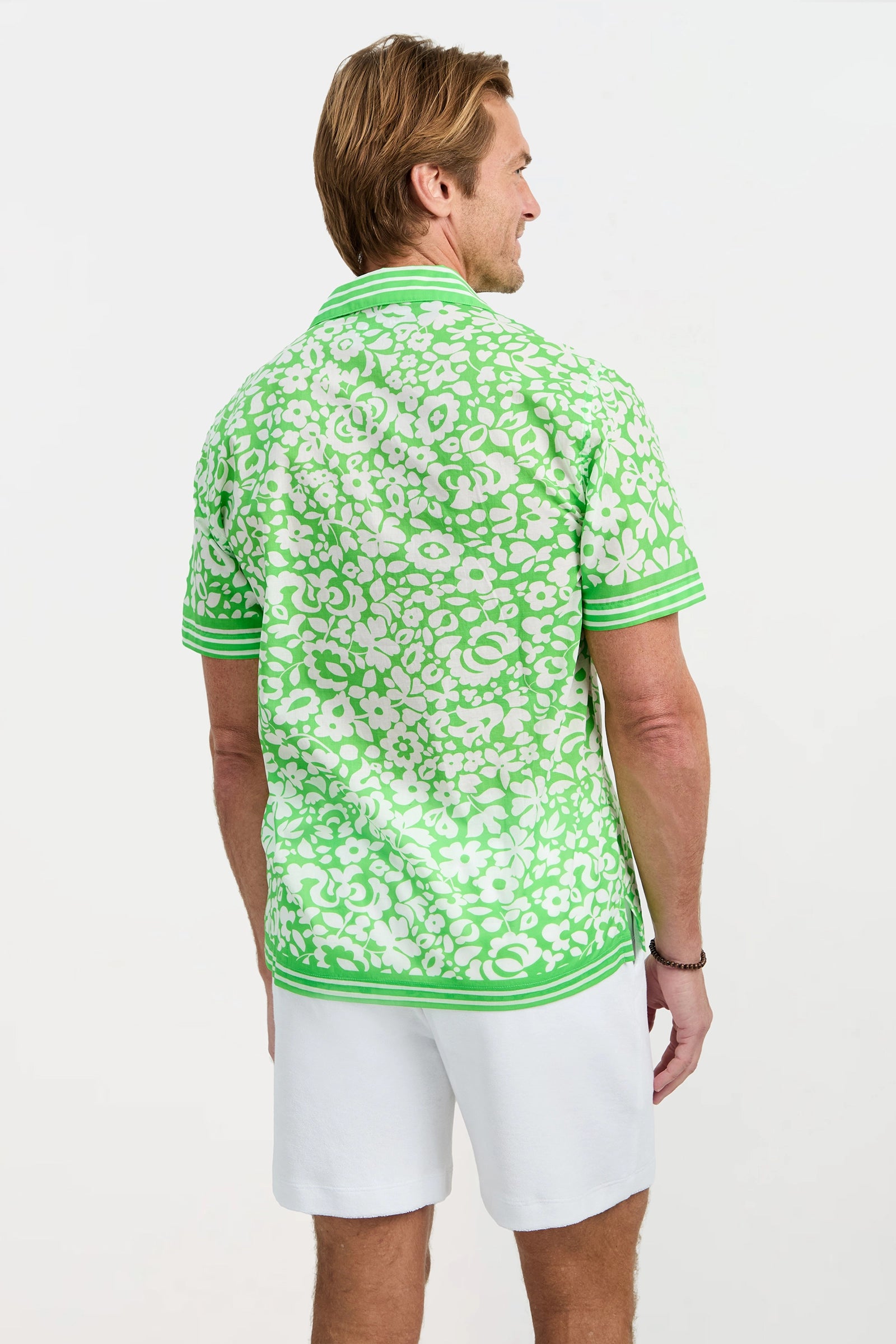 A man with light brown hair faces away, wearing the Josue Short-Sleeve Shirt in Poolside Gossip—a green and white floral-patterned shirt—paired with white shorts, standing against a plain light background.