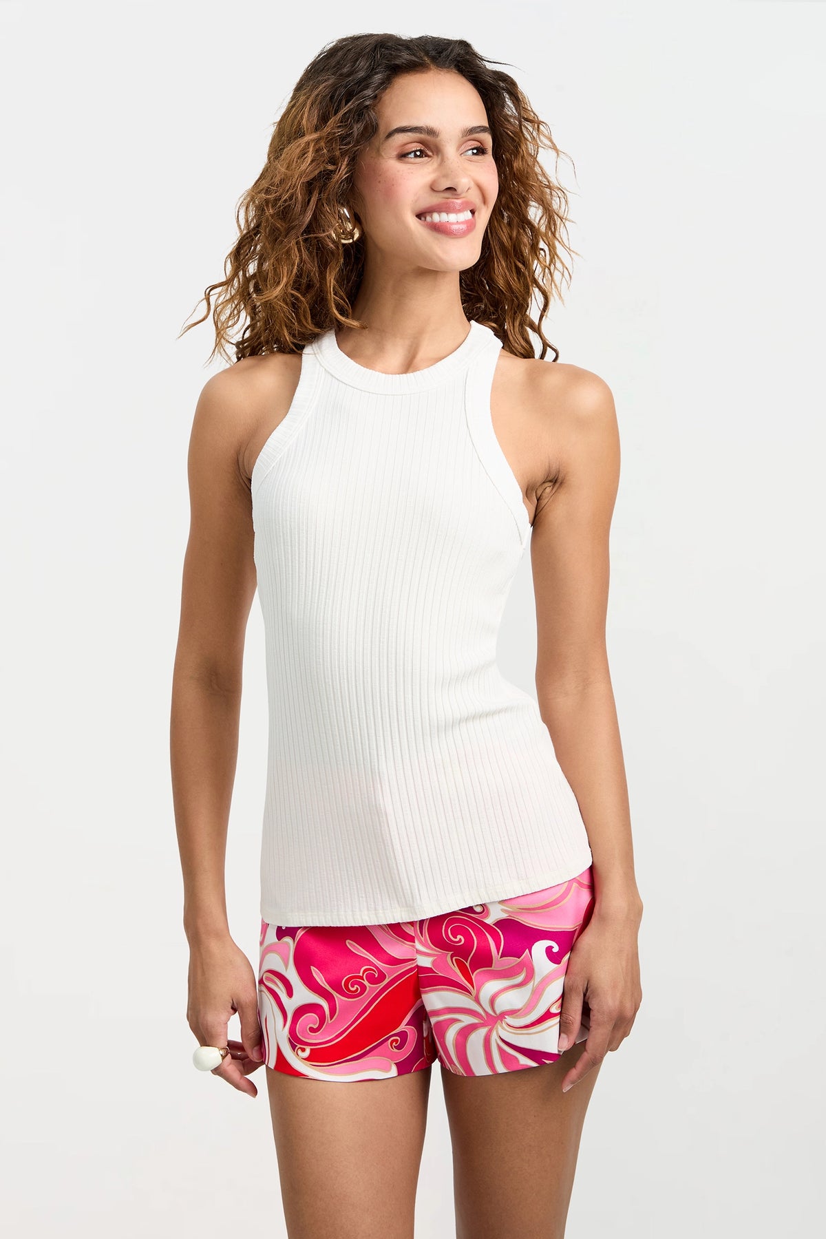 A woman with wavy brown hair smiles while wearing the Continental Tank Top, a sleeveless white ribbed style, paired with colorful pink and white patterned shorts against a plain light background.
