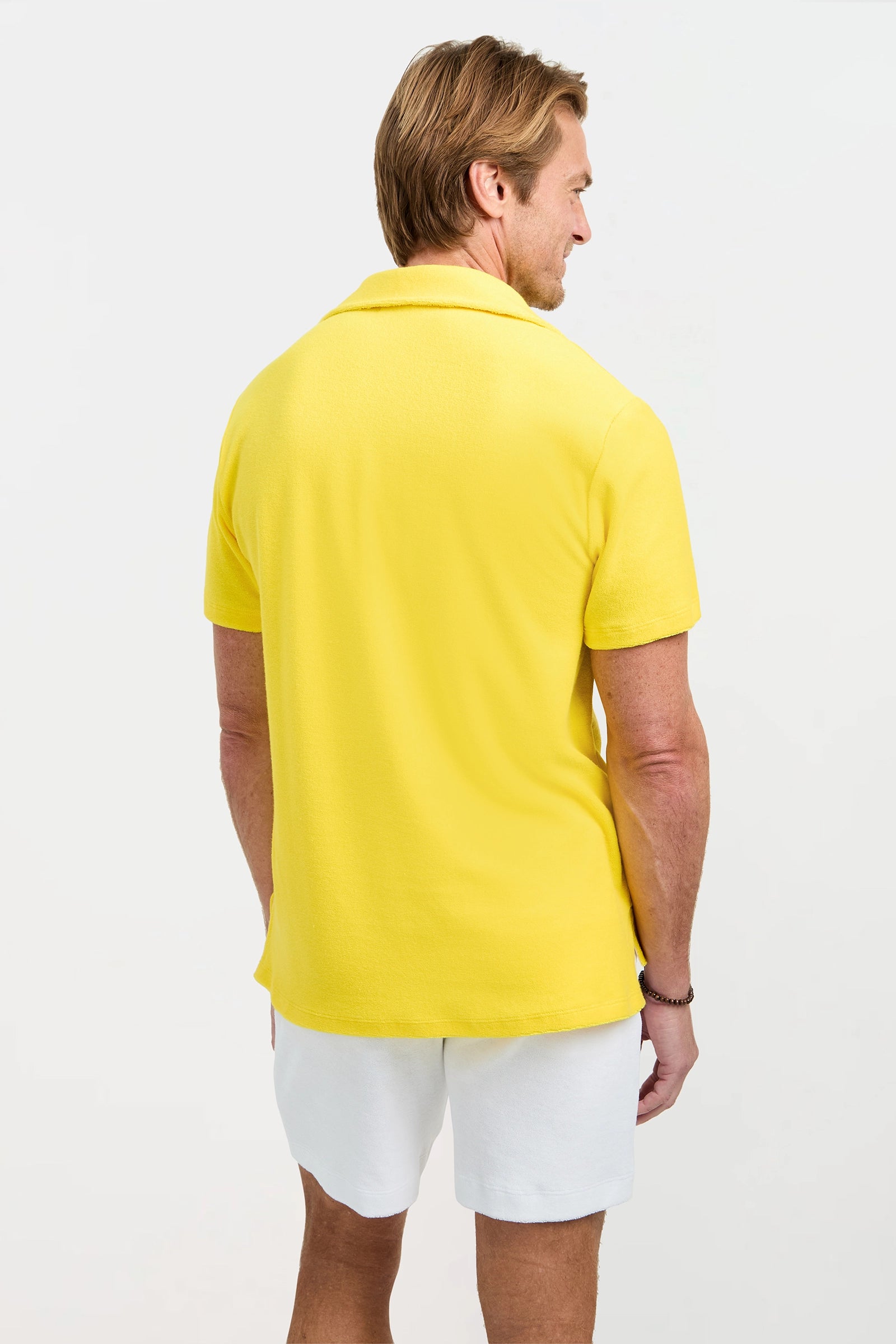 A man with light brown hair stands facing away in a bright yellow Dallas Short-Sleeve Polo Shirt - Linsk Terry and white shorts against a plain light background.