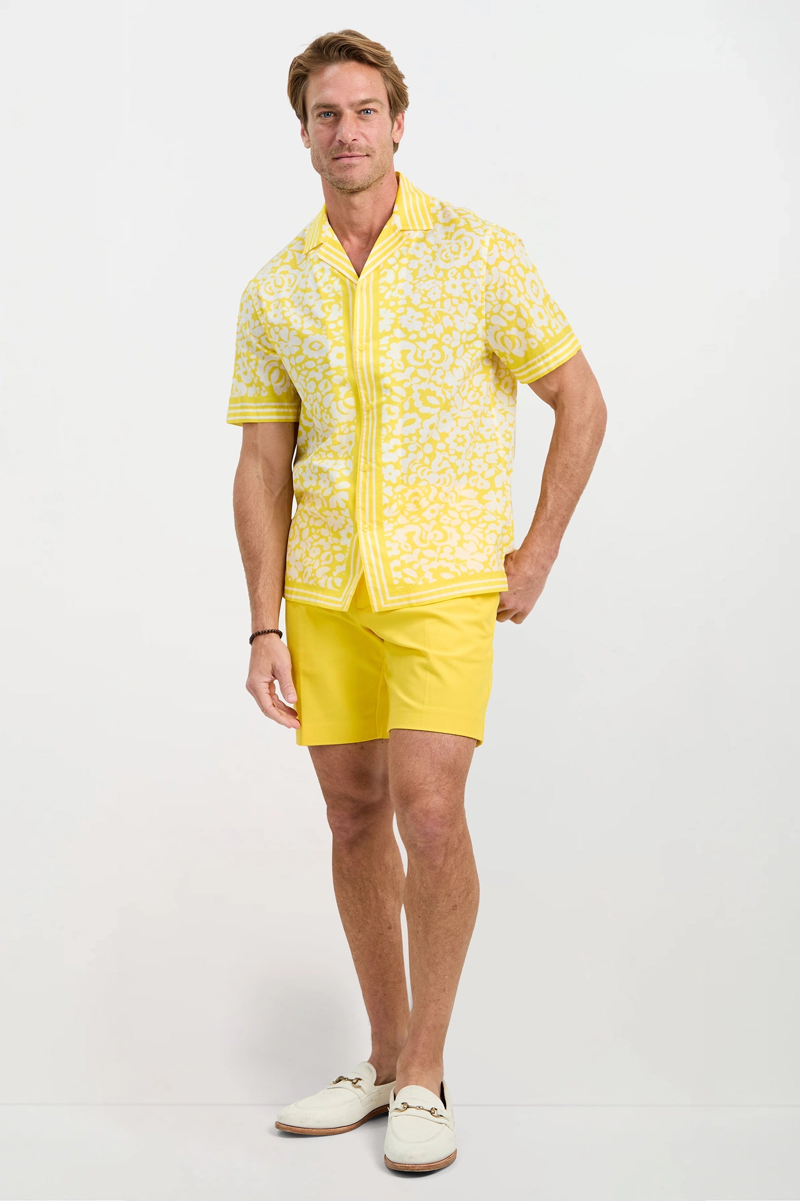 A man stands against a white background wearing the Josue Short-Sleeve Shirt in Poolside Gossip, yellow shorts, and white loafers. He smiles slightly with one hand in his pocket.
