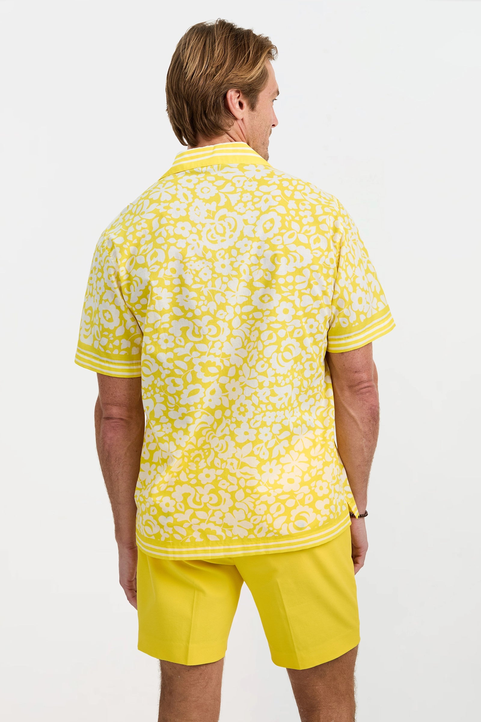 A man faces away from the camera wearing the Josue Short-Sleeve Shirt in Poolside Gossip, featuring a yellow and white floral pattern with matching shorts, set against a plain white background.
