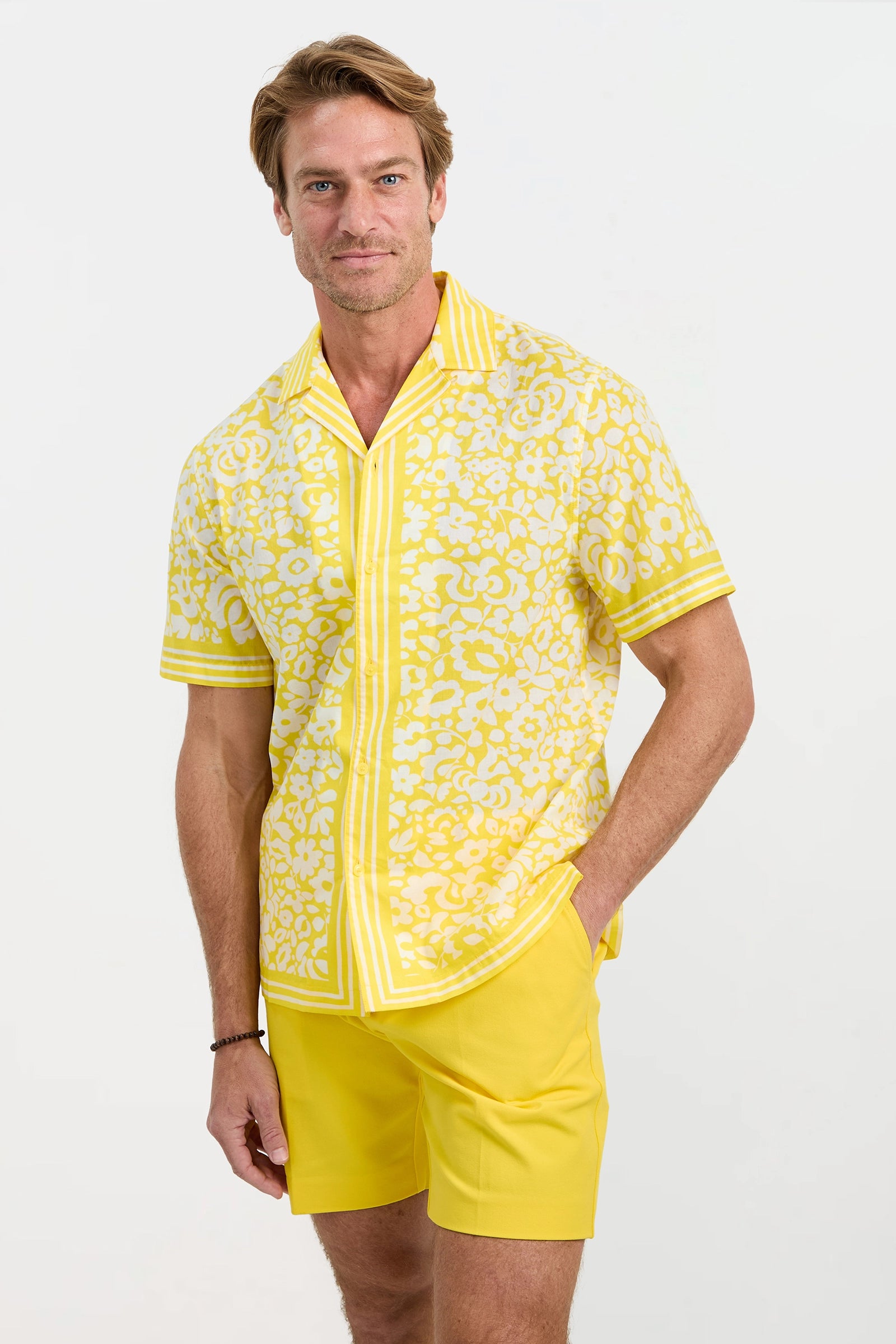 A man stands with one hand in his pocket, wearing the Josue Short-Sleeve Shirt in Poolside Gossip—a yellow and white floral-patterned shirt—paired with matching yellow shorts against a plain light background.