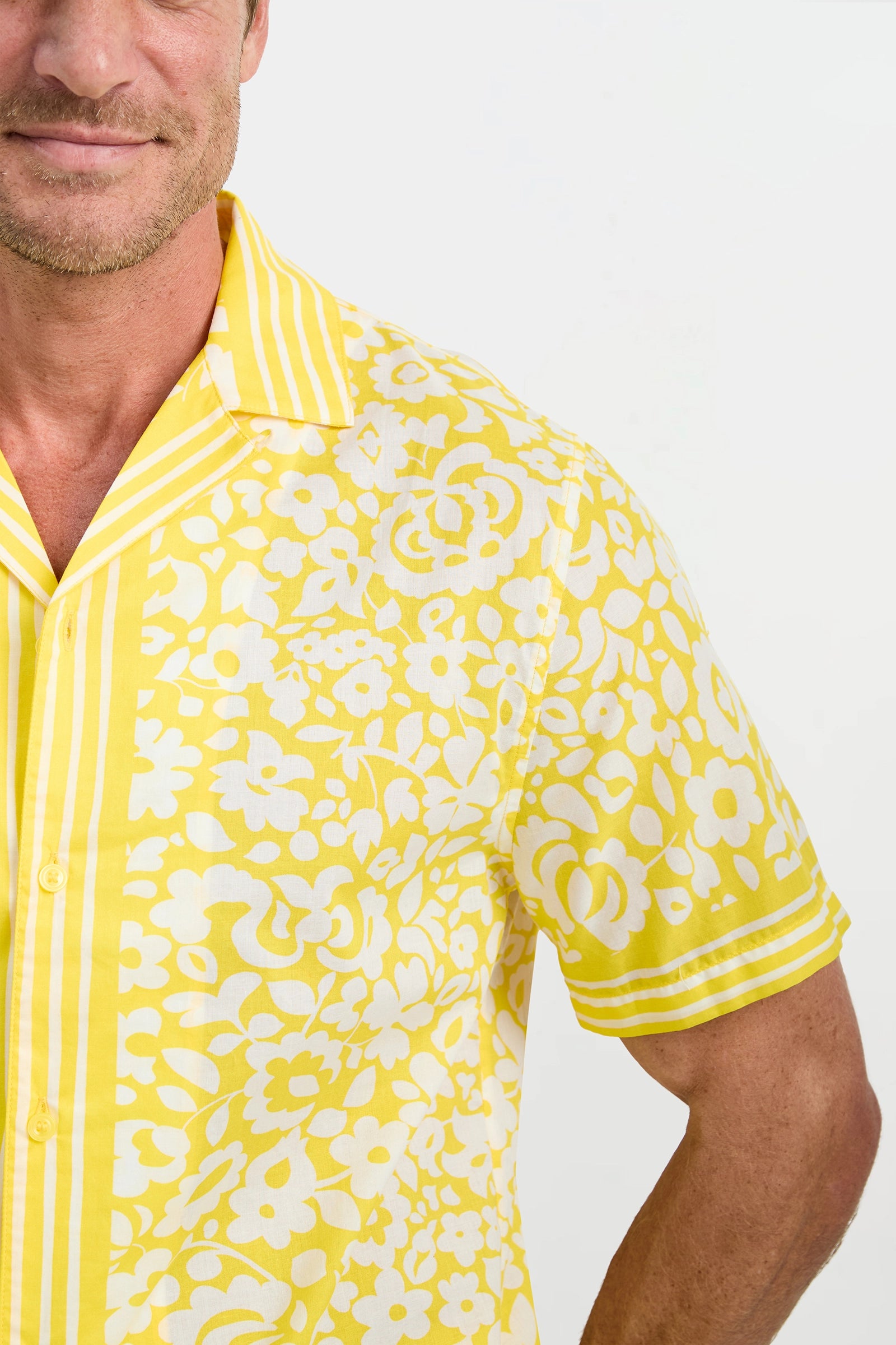 A man wears the Josue Short-Sleeve Shirt - Poolside Gossip, featuring yellow and white floral print and vertical stripes on the collar, button seam, and sleeves. Only the left side of his face and upper body appear against a plain background.