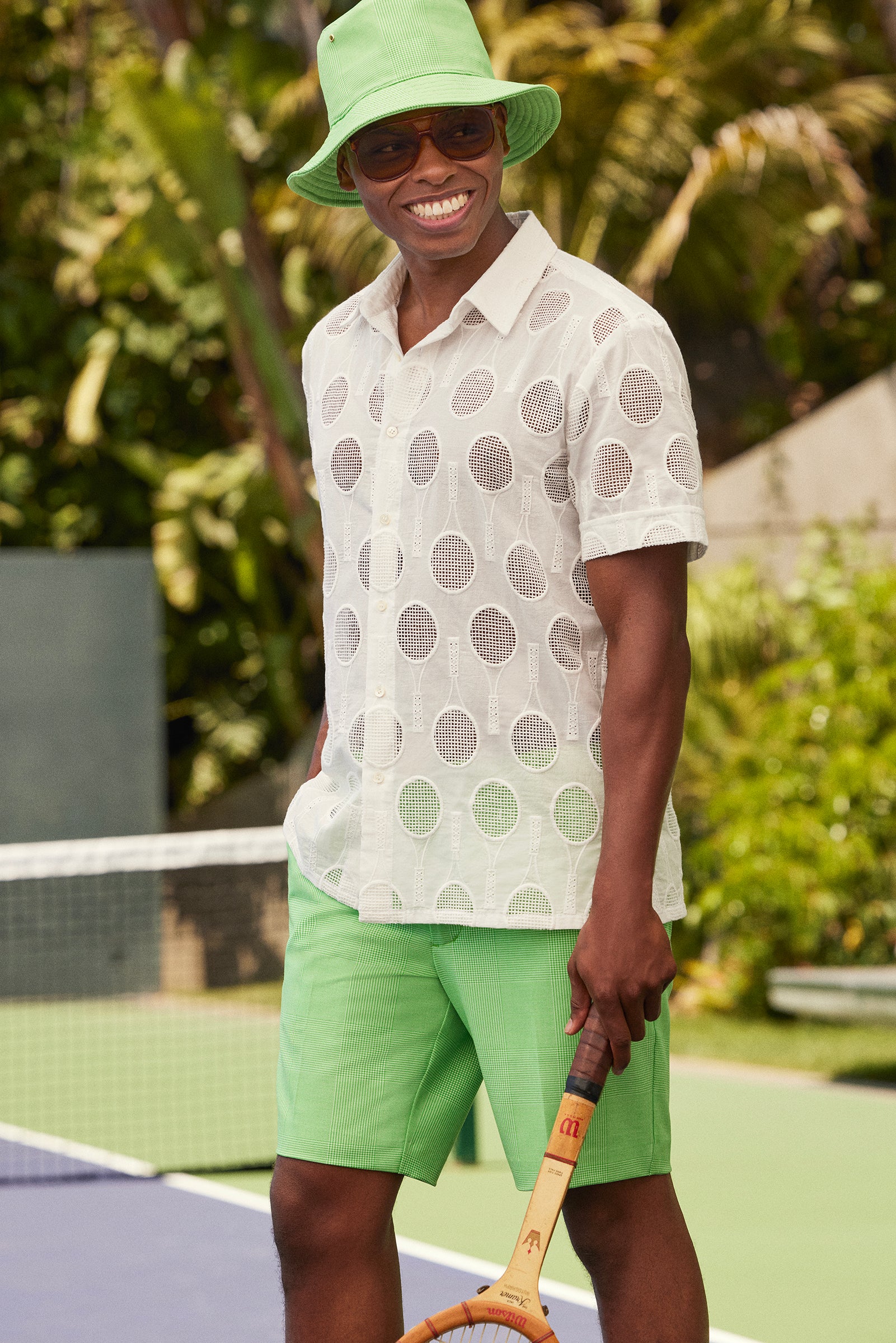 A smiling man on a tennis court wears sunglasses, a light green bucket hat, sheer white polka dot shirt, and the Travis Short - Game Time Glen Plaid, holding a wooden racket. Lush greenery and a net appear in the background.