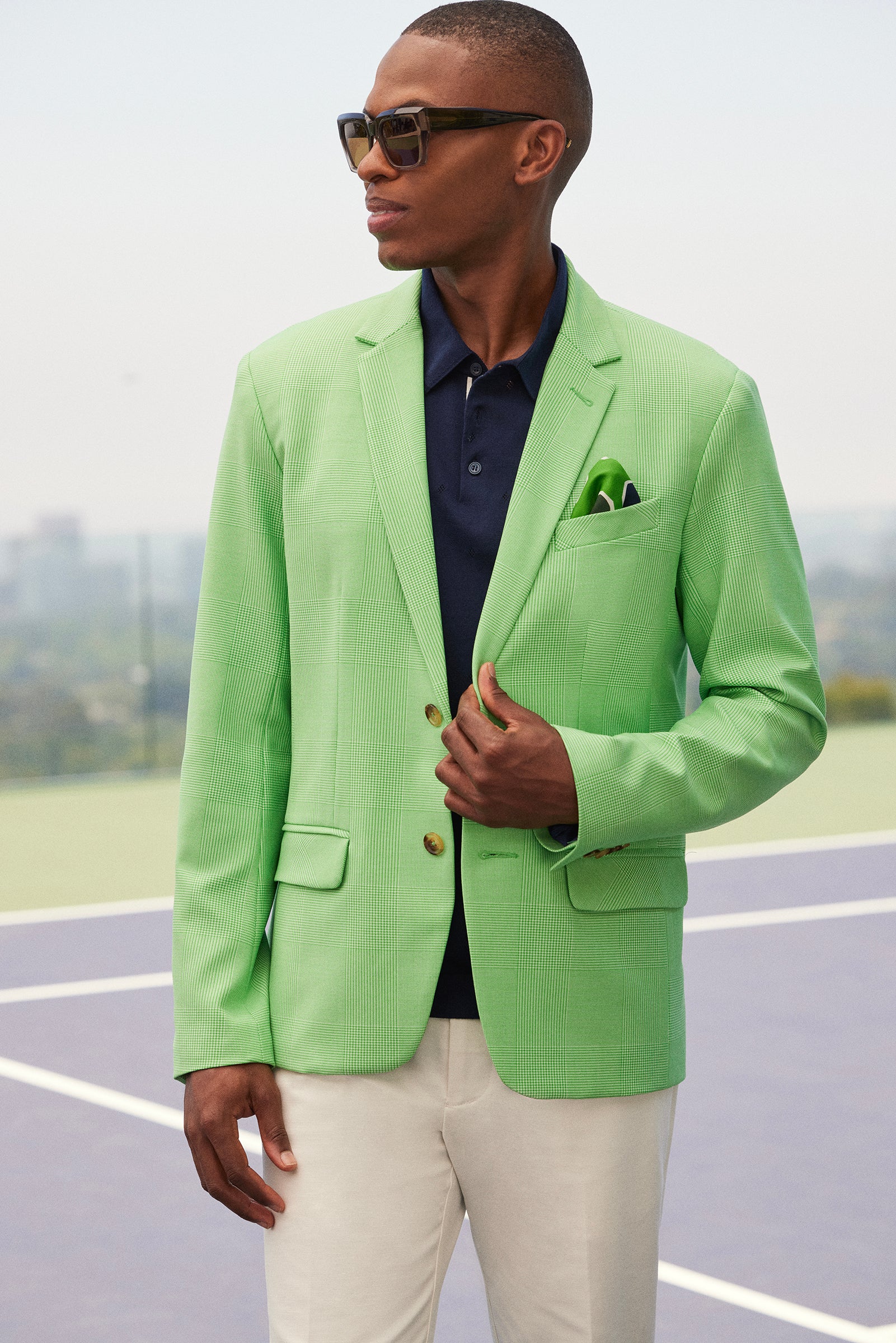 A man stands outdoors on a tennis court, wearing sunglasses, light-colored pants, and the Thurston Blazer - Game Time Glen Plaid over a navy polo shirt, looking to the side with one hand on his blazer.