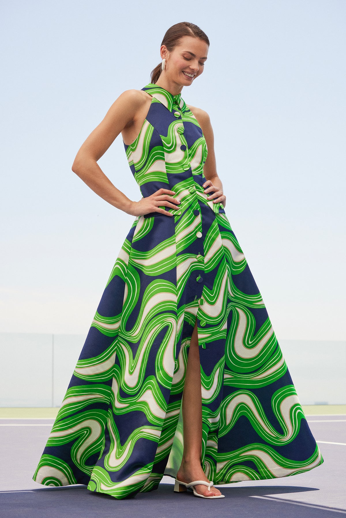 A woman wearing the Official Sleeveless Maxi Dress - Serena Satin, featuring a high neck, front buttons, and a bold green, blue, and white swirling pattern, stands outdoors smiling with her hands on her hips.