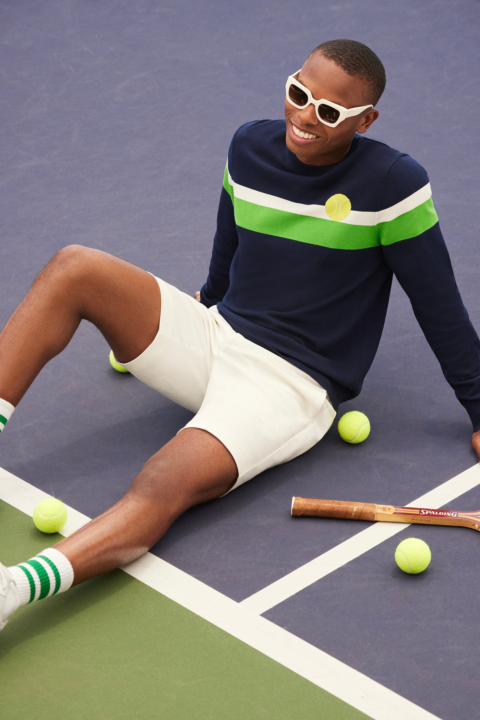 A man wearing the Romero Crewneck Sweater—navy with green and white stripes—and white shorts sits on a tennis court, smiling. He has sunglasses, striped socks, tennis balls, and a wooden racket around him.