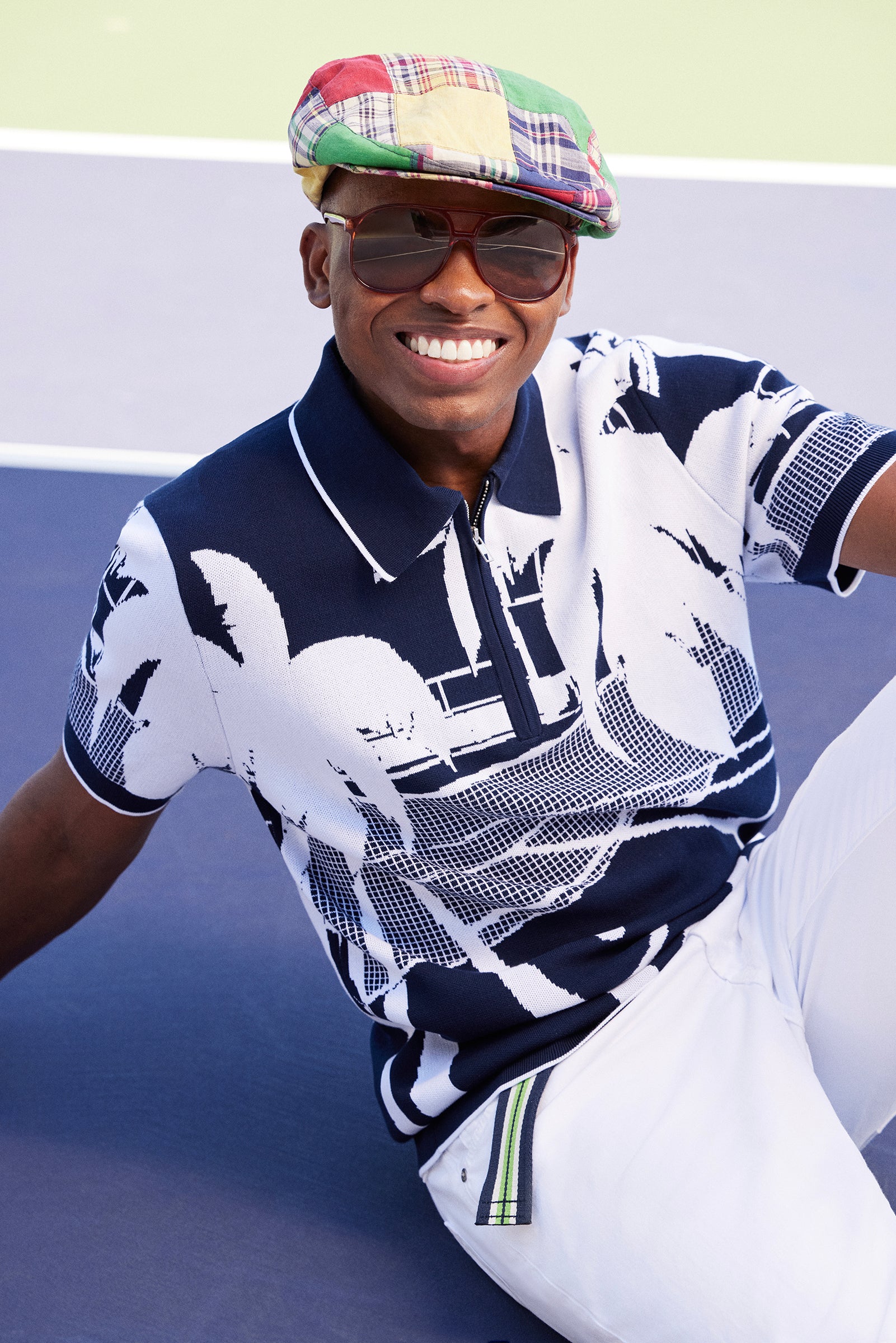 A smiling person wears the Grayson Short-Sleeve Polo - Graphic Knit, a colorful plaid cap, oversized sunglasses, and white pants while seated on a tennis court.