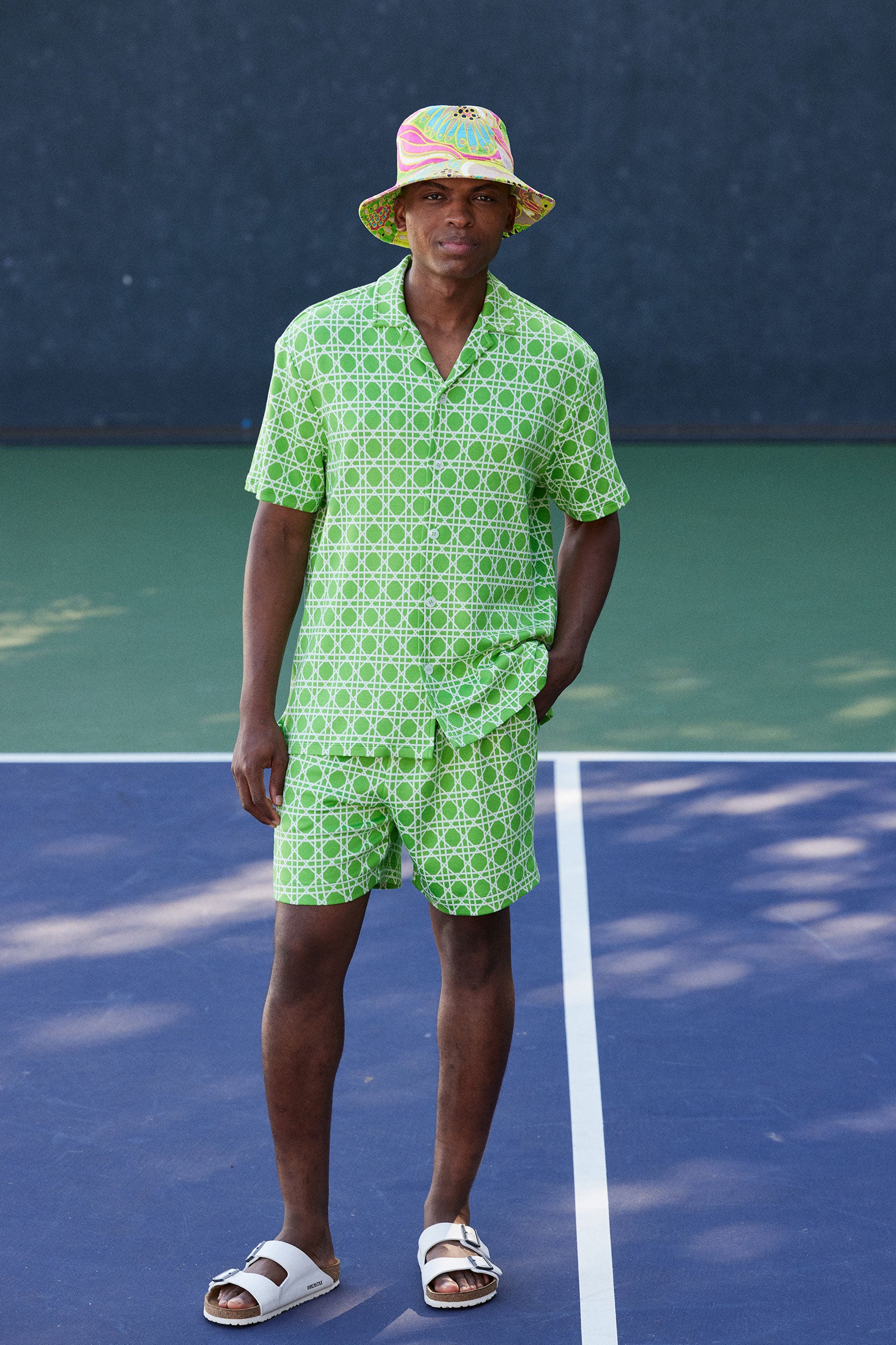 A man stands on a tennis court wearing the Lounge Short - Billie Jean, a green patterned shirt, a colorful bucket hat, and white sandals.