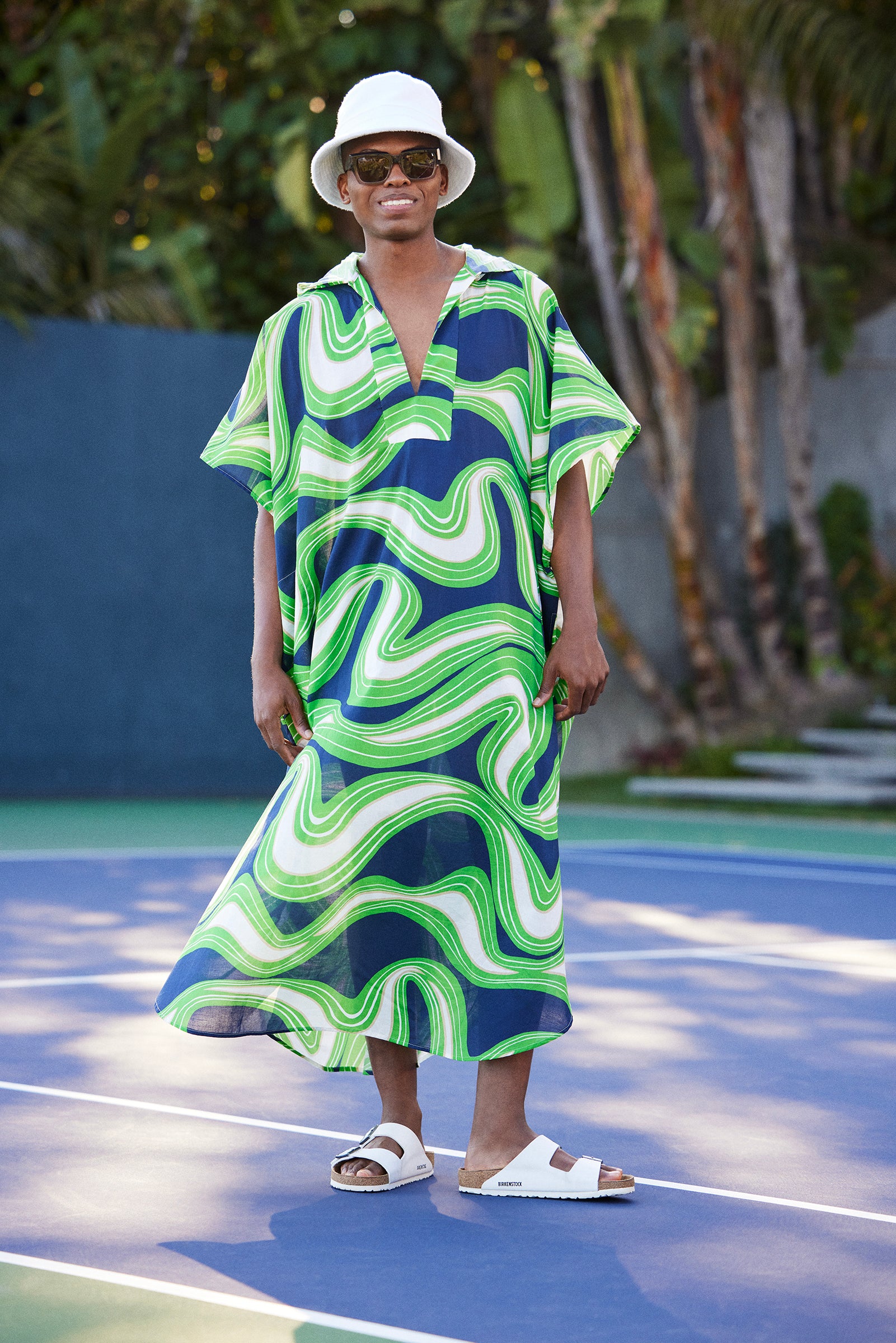 On a tennis court, someone wears the Hurley 2 Caftan - Serena Swirl, a flowing dress with green, white, and navy-blue waves, paired with a white bucket hat, sunglasses, and white sandals. Palm trees and greenery are in the background.