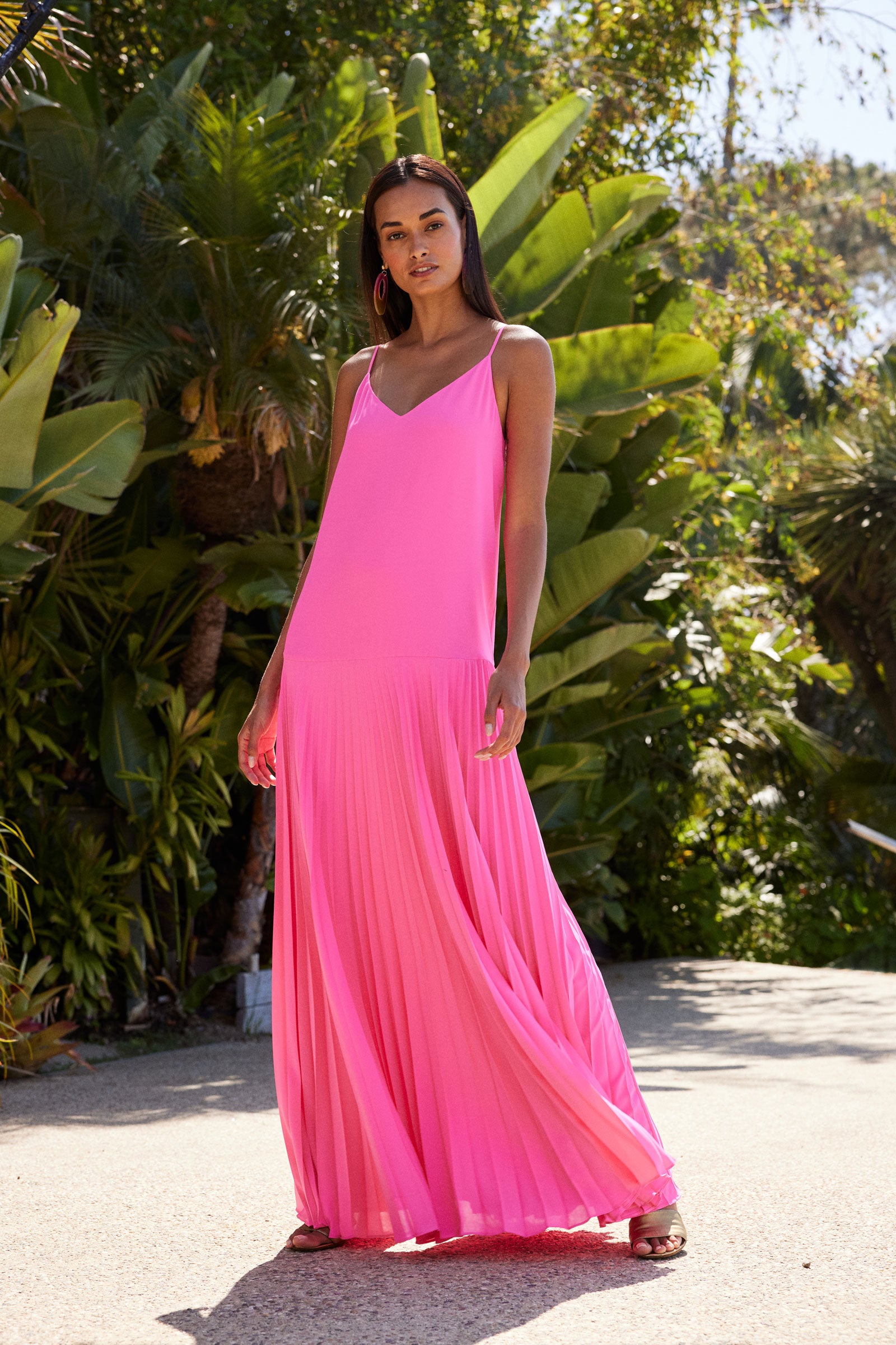 A woman stands outdoors on a sunny day wearing the Carve Sleeveless Drop-Waist Pleated Maxi Dress in bright pink. Lush green tropical plants and trees fill the background.