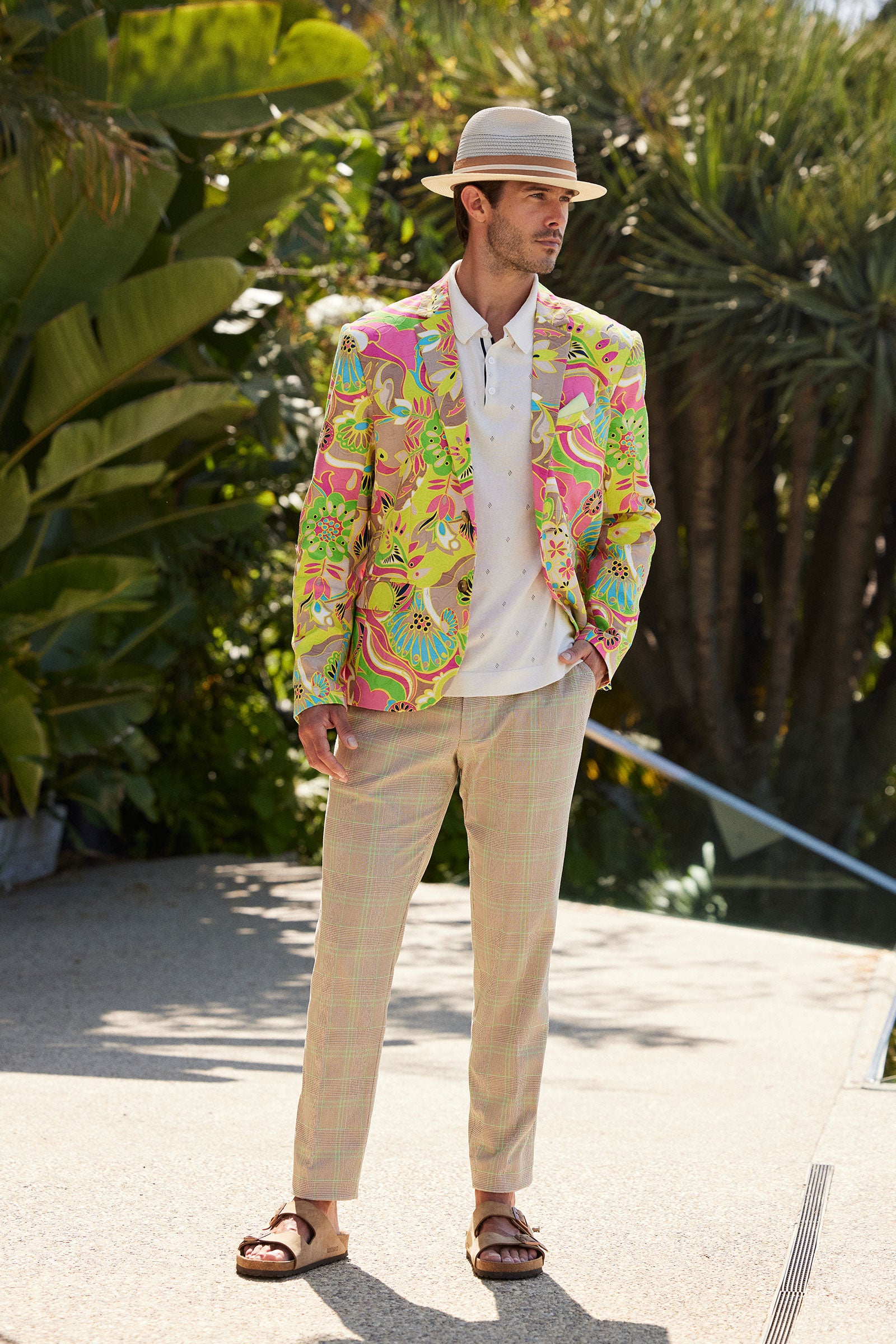 A man in the Thurston Blazer - Venus, paired with a white shirt, light plaid pants, brown sandals, and a beige fedora, stands outdoors near vibrant greenery on a sunny day.