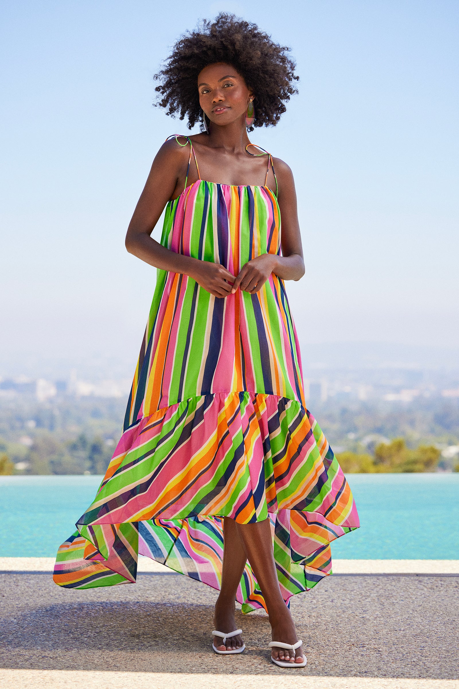 A woman with natural curly hair stands by a pool, wearing the Enchant 2 Sleeveless Dress in Splash Stripe with thin straps and white sandals. She holds her hands together, cityscape and sky visible in the background.