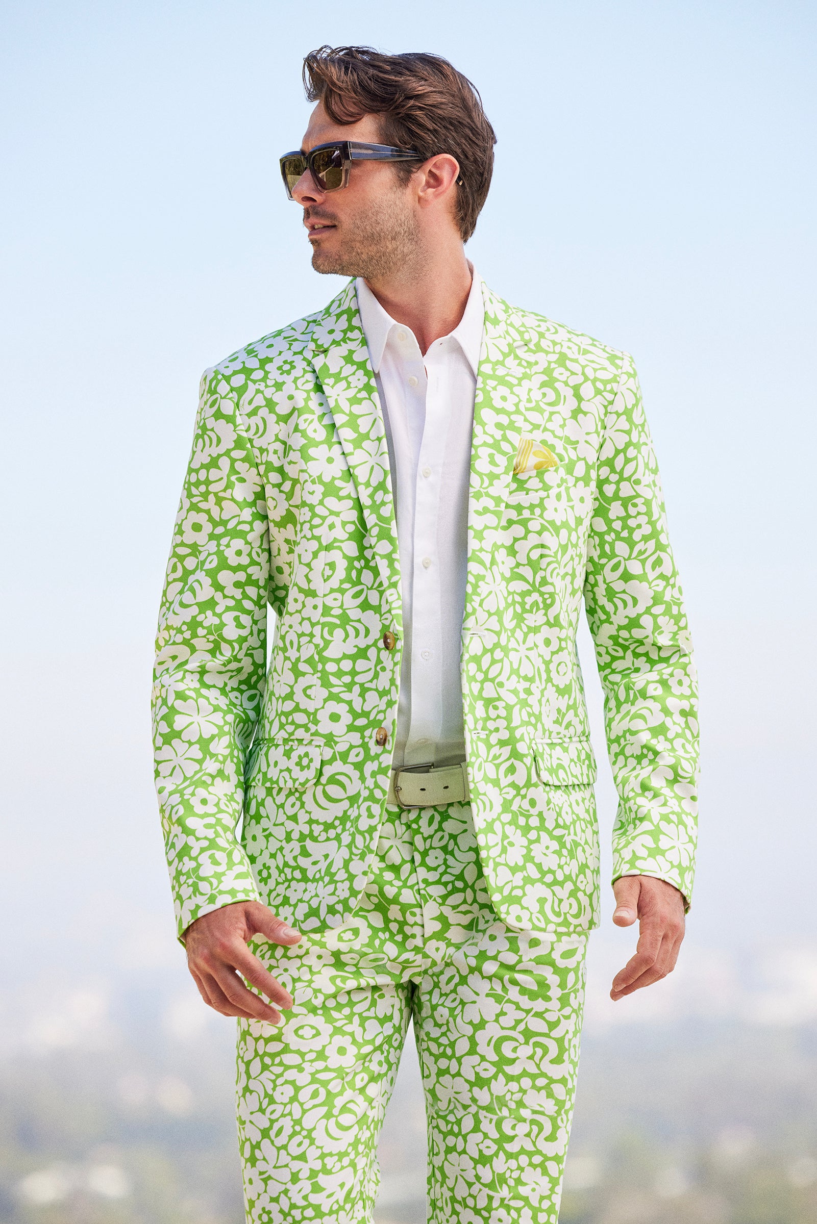 A man in sunglasses and a white shirt wears the Thurston Blazer - Poolside Gossip, a bright green and white floral-patterned suit, while standing outside against a softly blurred blue sky.