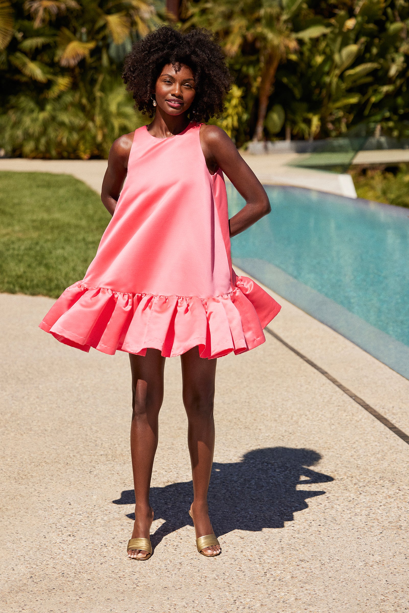 A woman poses by a pool in the Winner Sleeveless A-Line Dress - Solid Satin, a bright pink ruffled style, paired with gold open-toe heels. She smiles with her hands behind her back amid lush greenery.