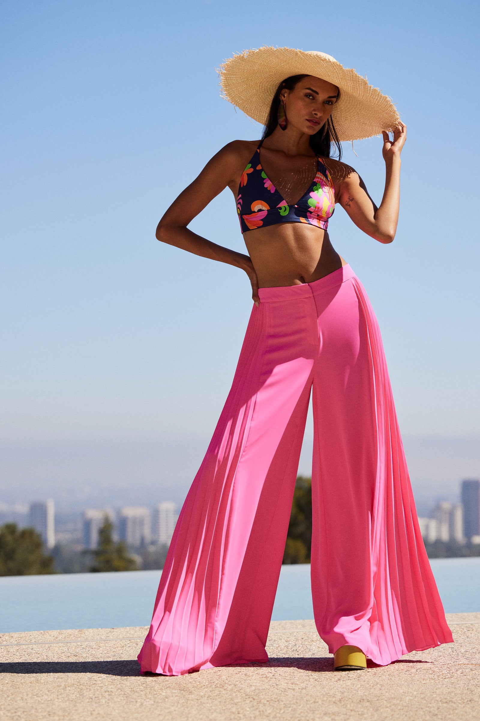 A woman poses outdoors in Frame Wide-Leg Pleated Pant - GS Georgette, paired with a floral bikini top and sunhat, set against a city skyline and blue sky.