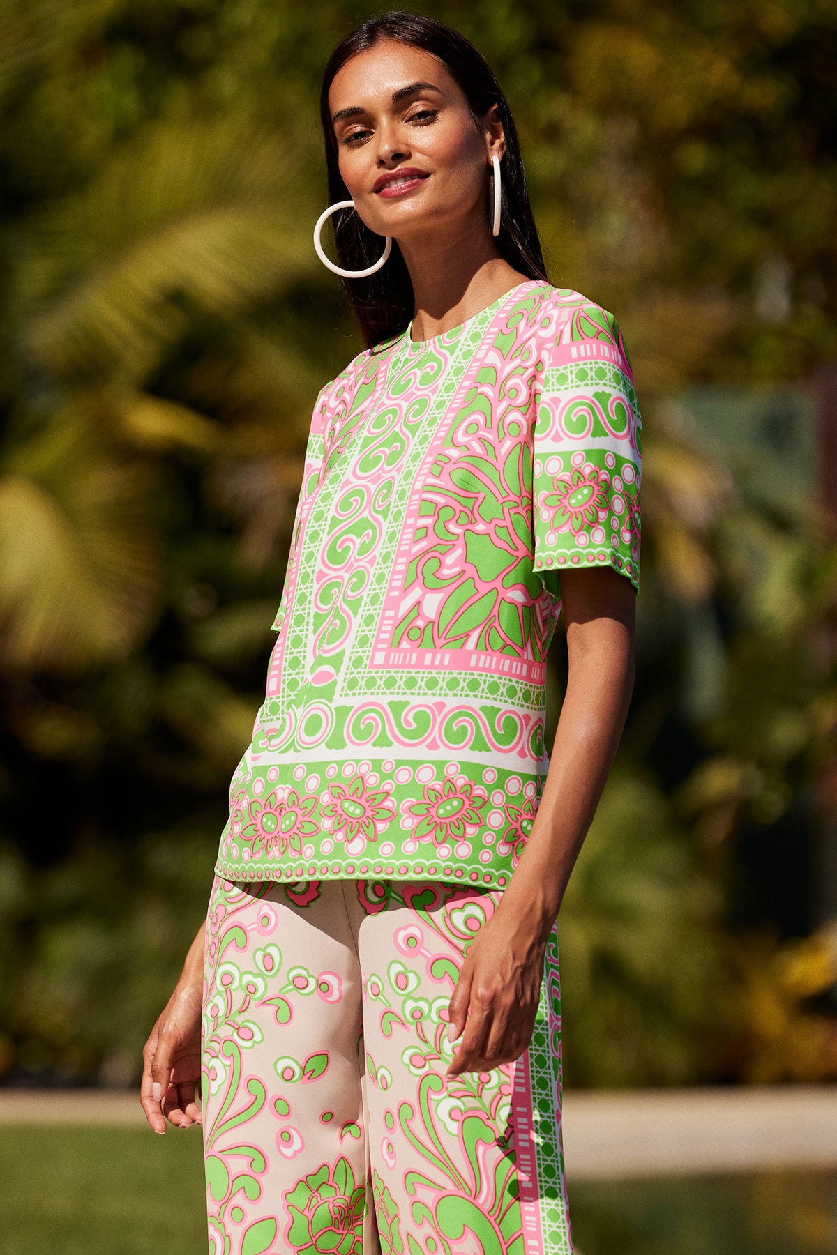 A woman with long dark hair and hoop earrings stands outdoors, smiling slightly as she wears the Riggs Short-Sleeve Top in Petra Floral, a matching set featuring bold pink, green, and white floral and geometric patterns. Greenery is behind her.