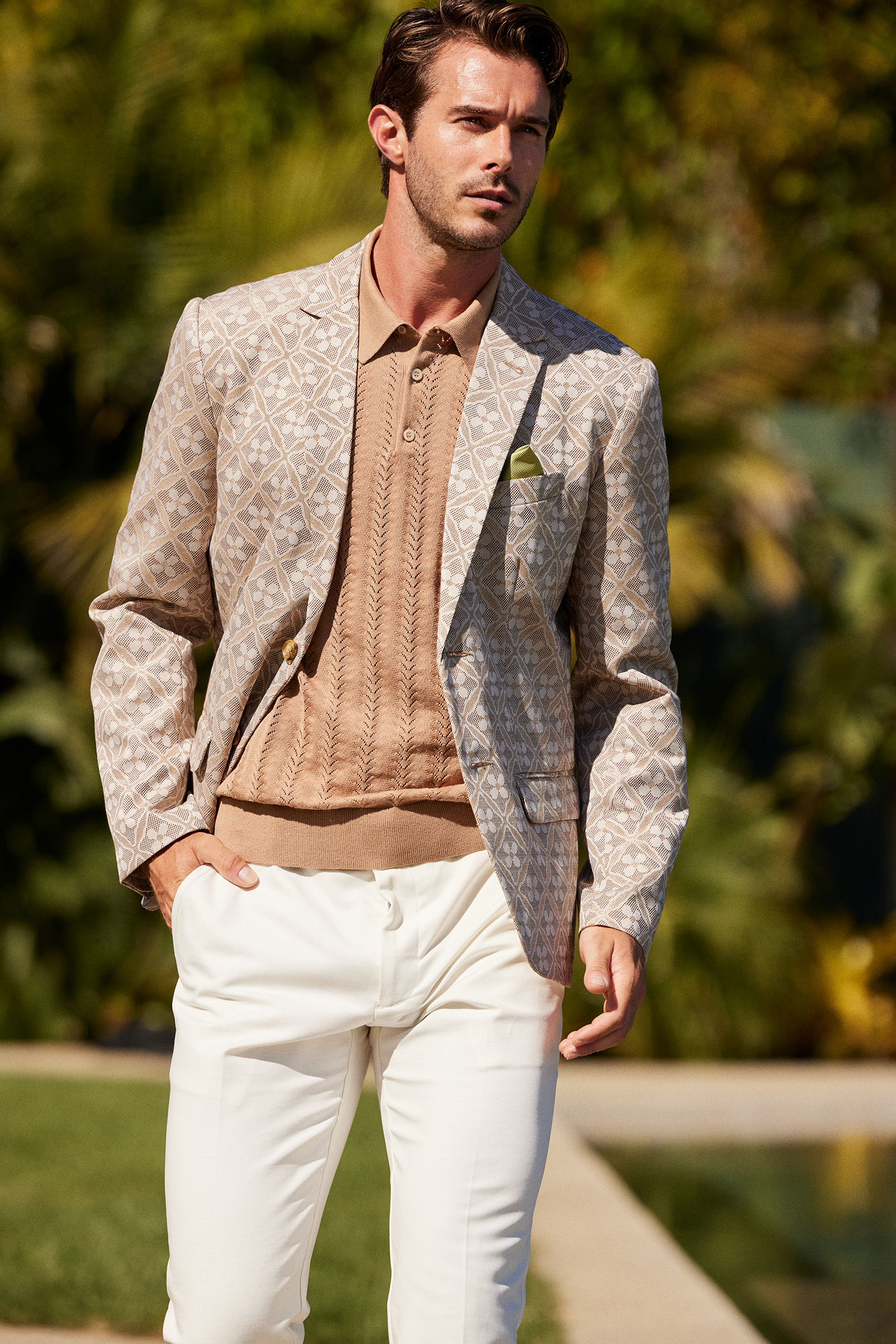 A man stands outdoors with his hand in his pocket, surrounded by greenery, wearing the Thurston Blazer - Metallic Daisy Jacquard, a tan knit polo shirt, and white pants.