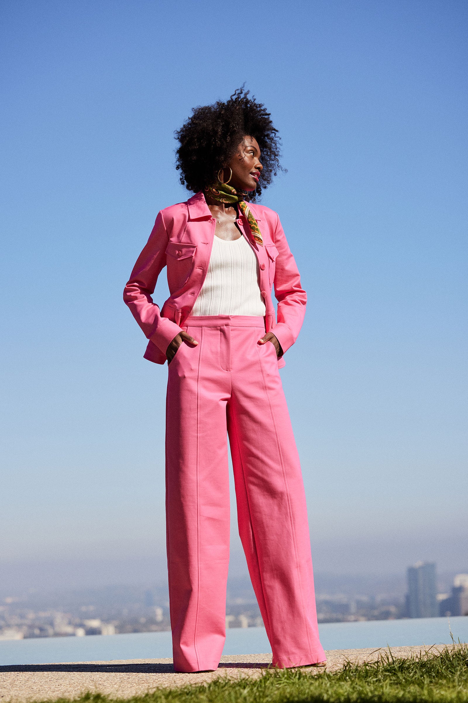 A woman with curly hair stands outdoors against a city skyline, wearing the Half Court Wide-Leg Pant -Solid Suiting in bright pink, paired with a white ribbed top and a patterned scarf.