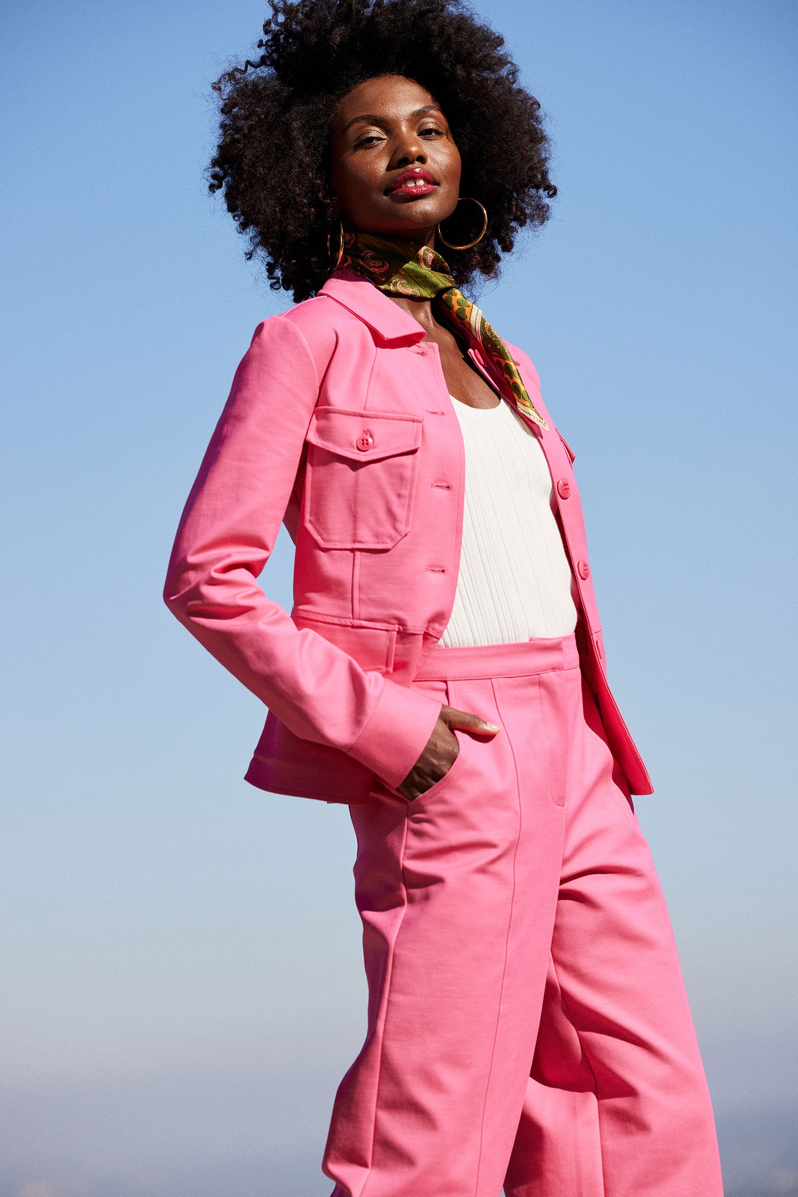A woman with natural curly hair stands confidently outdoors under a clear blue sky, wearing the Qualifier Button-Front Peplum Jacket - Solid Suiting in bright pink over a white top and a patterned scarf around her neck.