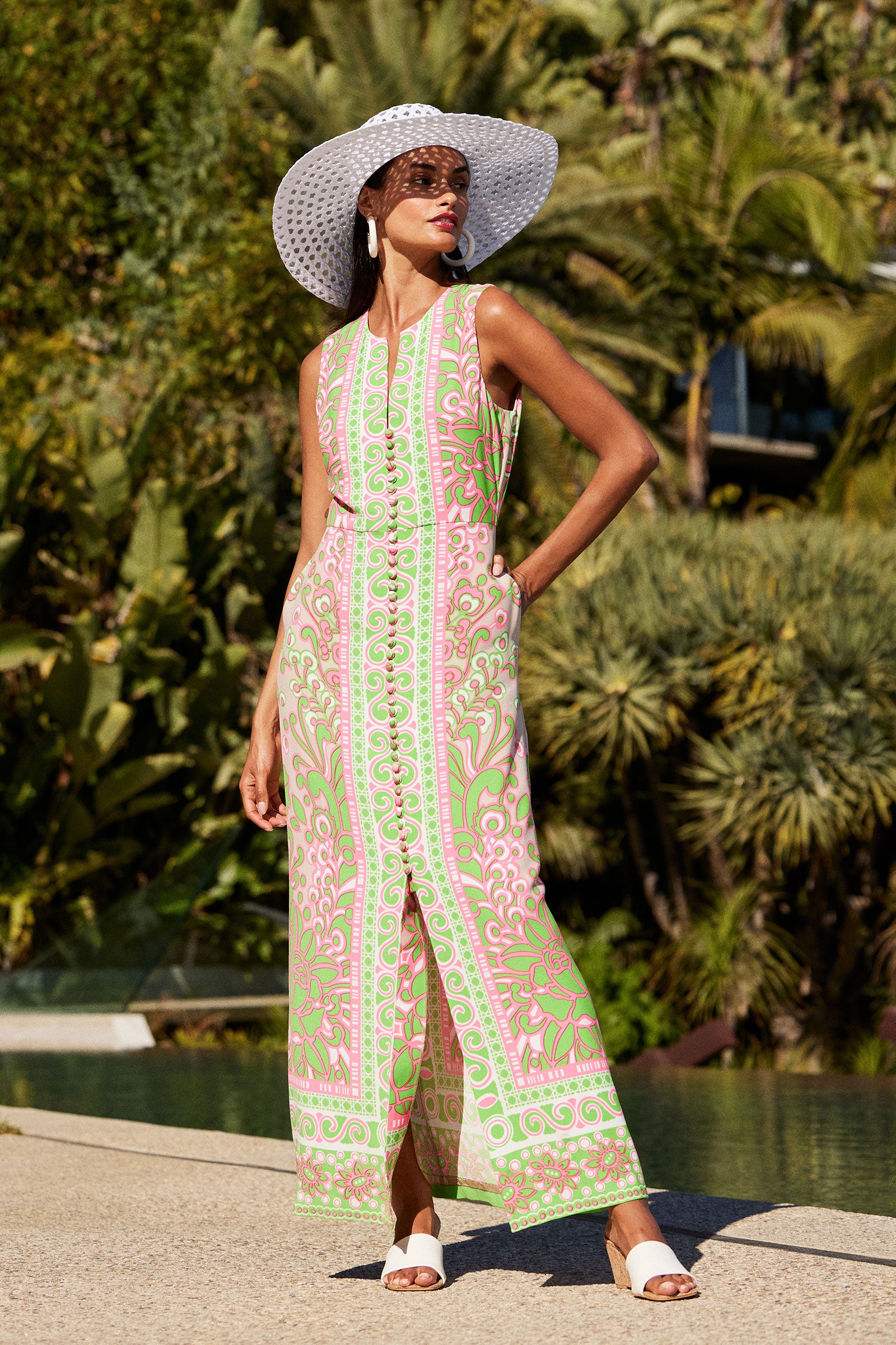 A woman stands by a pool in lush greenery, wearing the Novak Sleeveless Midi Dress - Petra Floral, styled with a wide-brimmed white hat, white earrings, and white slide sandals.