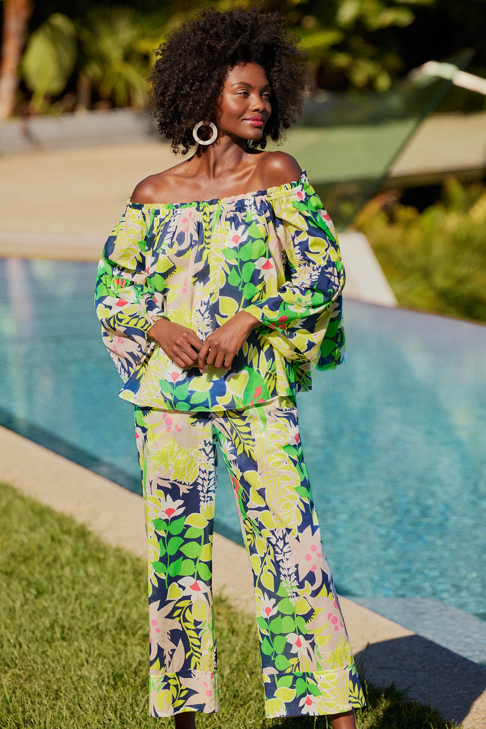 A woman with curly hair and large hoop earrings stands by a pool, smiling to the side in the Perry Off-Shoulder Top - PS Eternal, a long-sleeve piece featuring a vibrant tropical leaf print and matching pants.