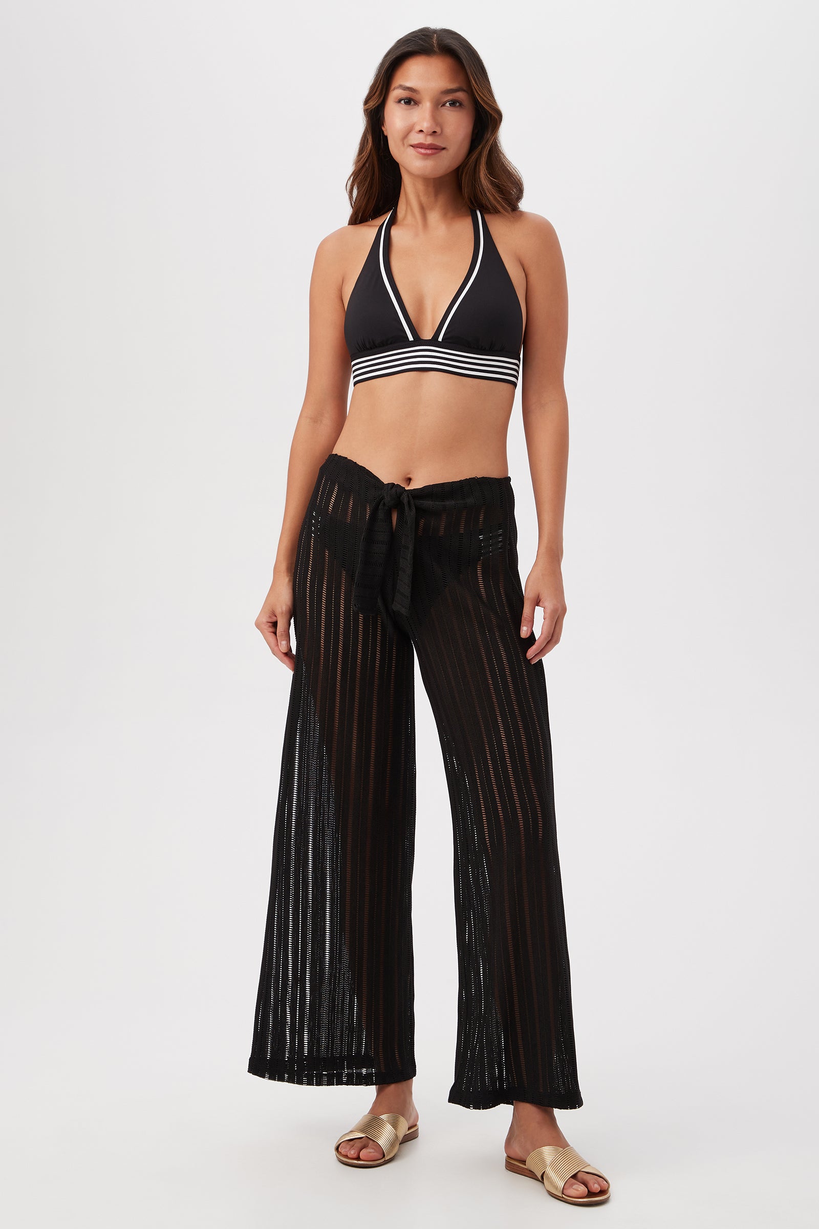 A woman models the Skyhigh Mesh Tie-Front Beach Pant Cover-Up over a black halter bikini top with white trim and gold slide sandals, creating a chic beach-ready look against a plain white background.