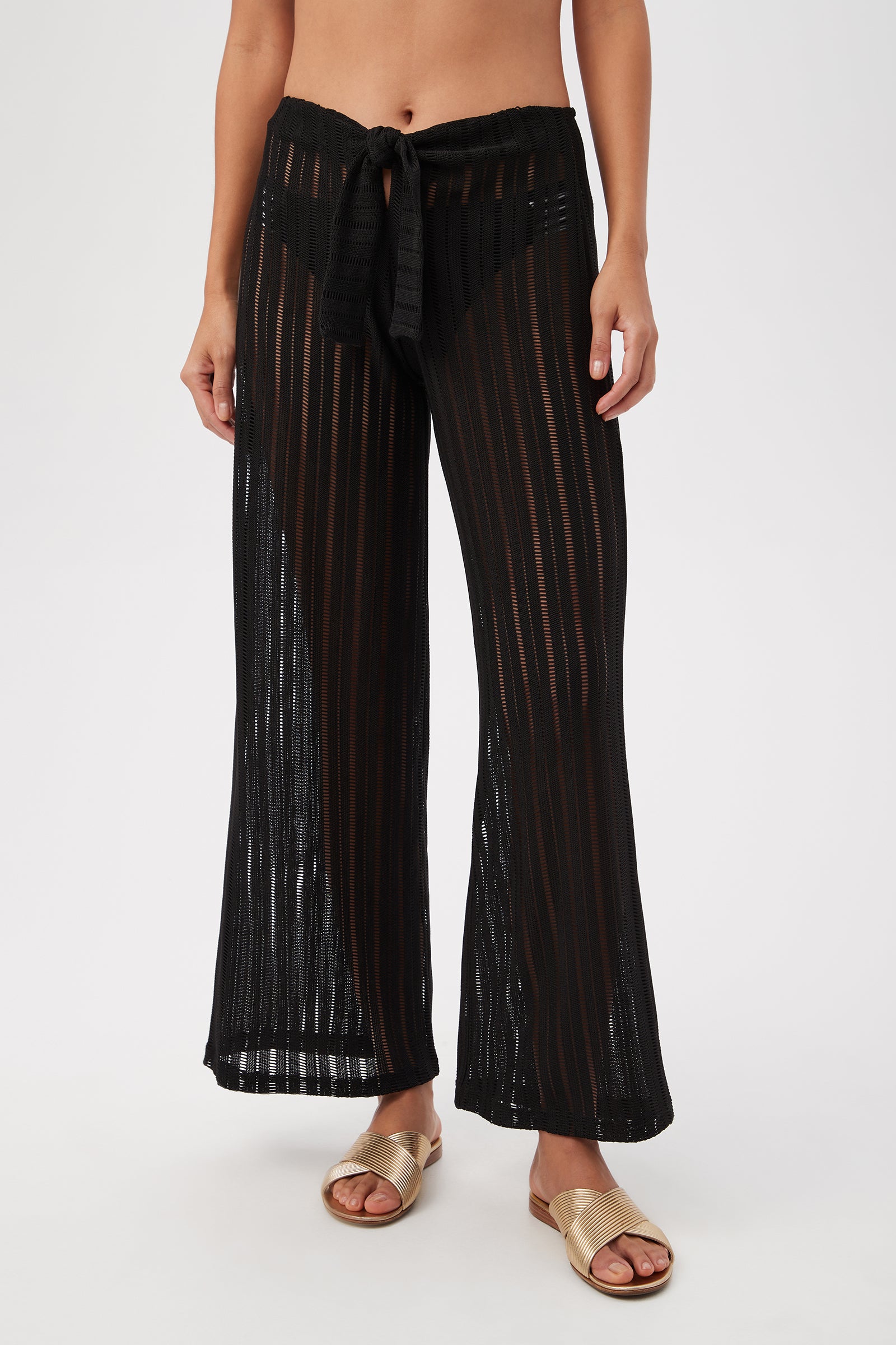 Someone is wearing the Skyhigh Mesh Tie-Front Beach Pant Cover-Up in black, featuring sheer wide-leg pants with vertical stripes, front waist tie, and revealing black underwear beneath. The outfit is styled with gold flat sandals. Torso not fully visible.