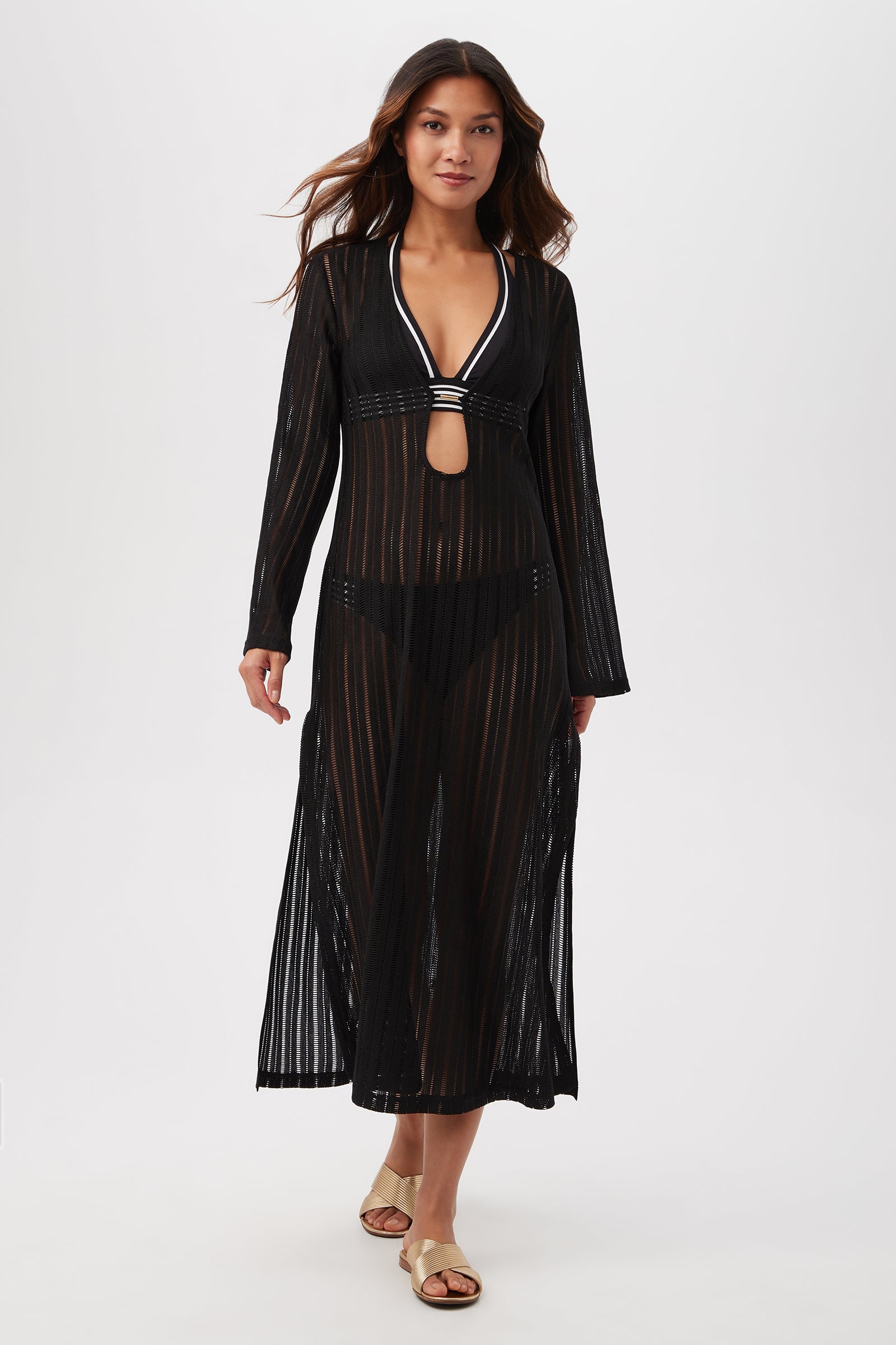 A woman walks forward against a plain white background wearing the black Skyhigh Long-Sleeve Mesh Maxi Dress Cover-Up over a black bikini, paired with flat sandals.
