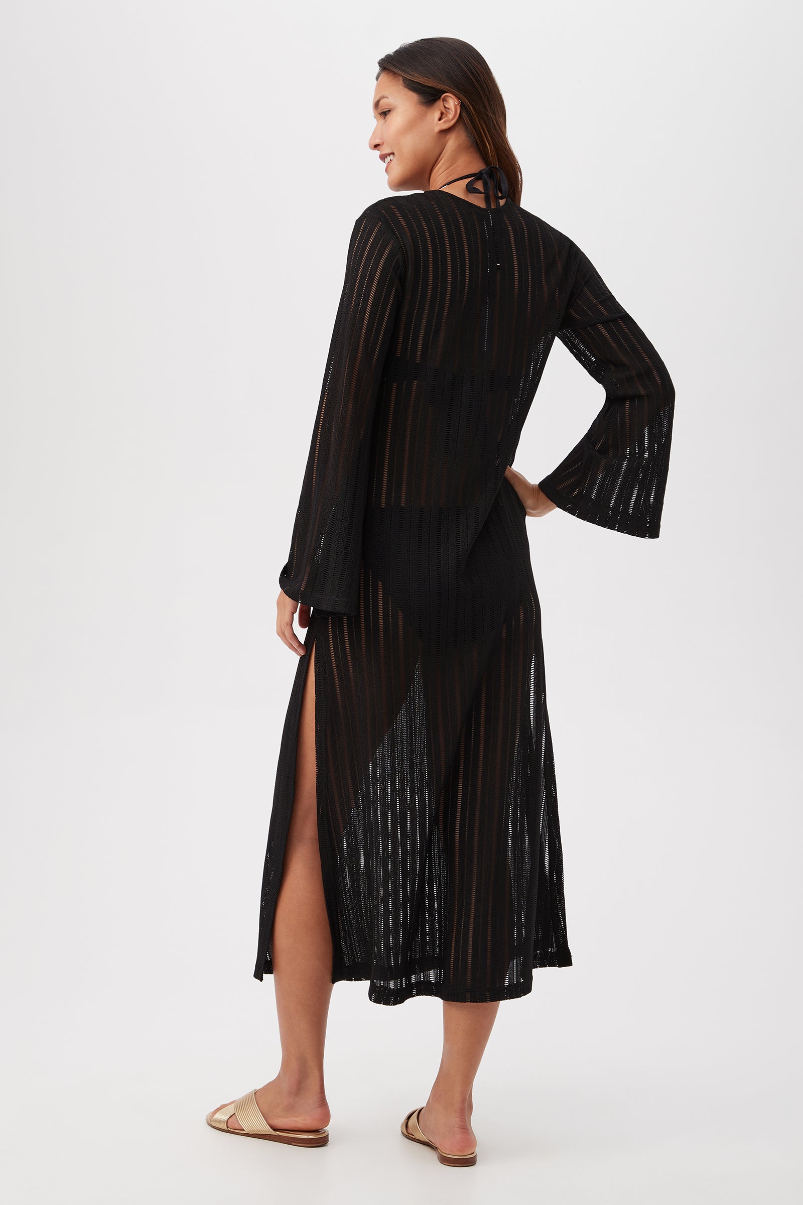 A woman with long hair, wearing tan flat sandals, smiles slightly as she faces away in the Skyhigh Long-Sleeve Mesh Maxi Dress Cover-Up over swimwear. The sheer black dress features vertical stripes against a plain white background.