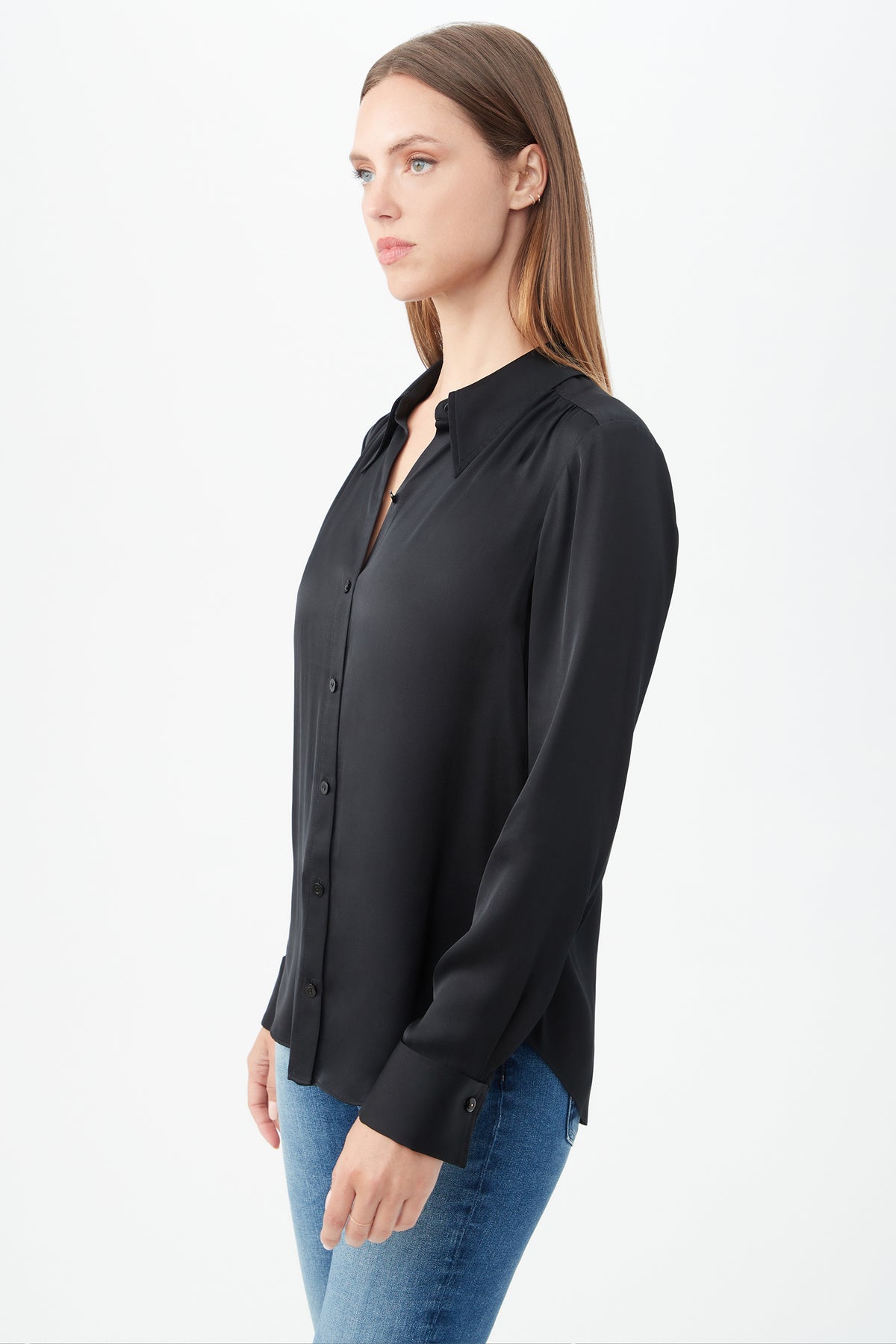 A woman with straight, light brown hair wears the Cassavetes Long-Sleeve Stretch Silk Button-Up Shirt in black and blue jeans, standing sideways against a white background to showcase effortless women's fashion.