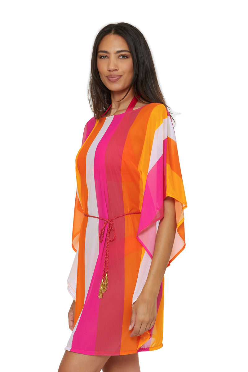 A woman models the Anissa Cover-Up Caftan - Sunburst, a colorful dress with bold orange, pink, red, and white vertical stripes, loose sleeves, and a thin belt, as she smiles softly against a white background.