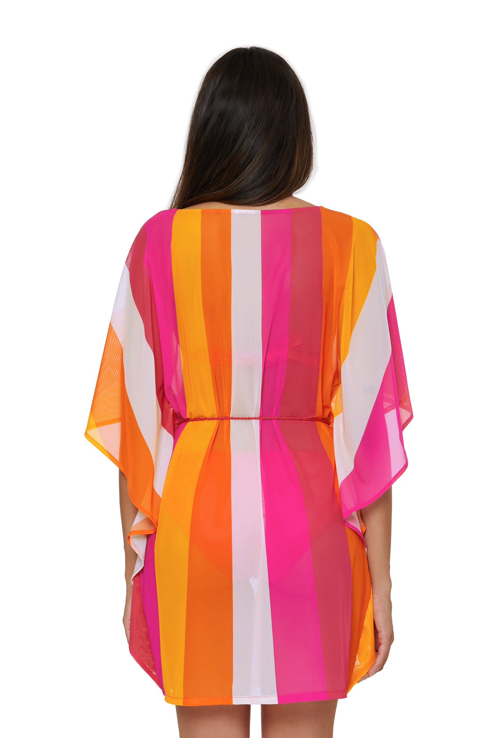 A woman with long brown hair, seen from behind, wears the Anissa Cover-Up Caftan in Sunburst—a short, sheer dress featuring wide vertical stripes in pink, orange, yellow, and white with loose sleeves and a cinched waist.