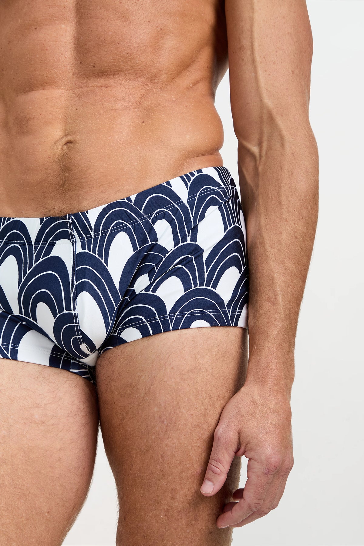 A close-up of a man wearing the Sorrento Swim Trunk - Arches, featuring a navy blue and white abstract semicircle pattern, with his bare torso and right arm visible against a neutral background.