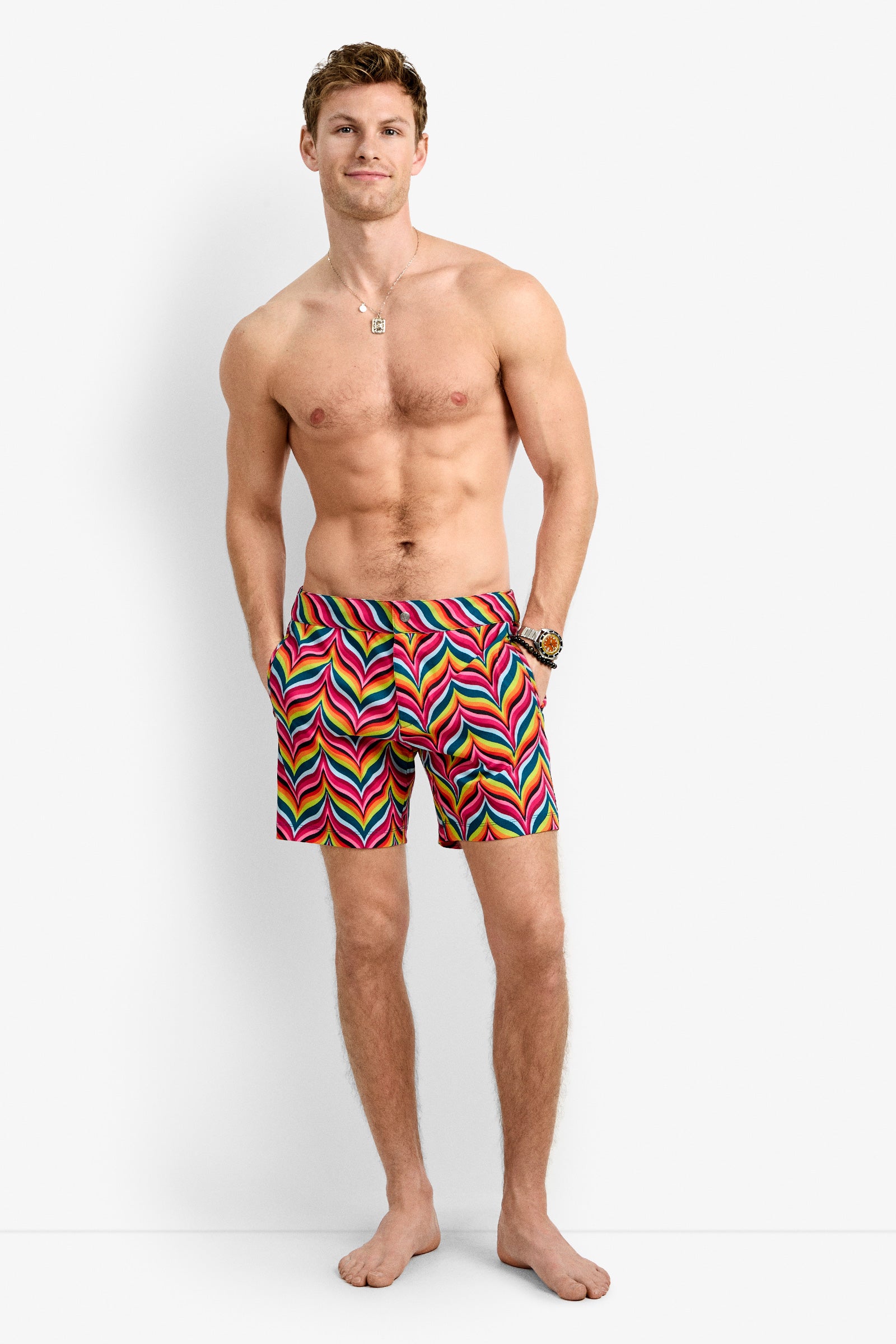 A shirtless man stands barefoot against a white background, smiling slightly with his hands in his pockets, wearing the Honolulu Board Short - Chelsea Chevron featuring a vibrant multicolored chevron pattern for stylish summer swimwear.