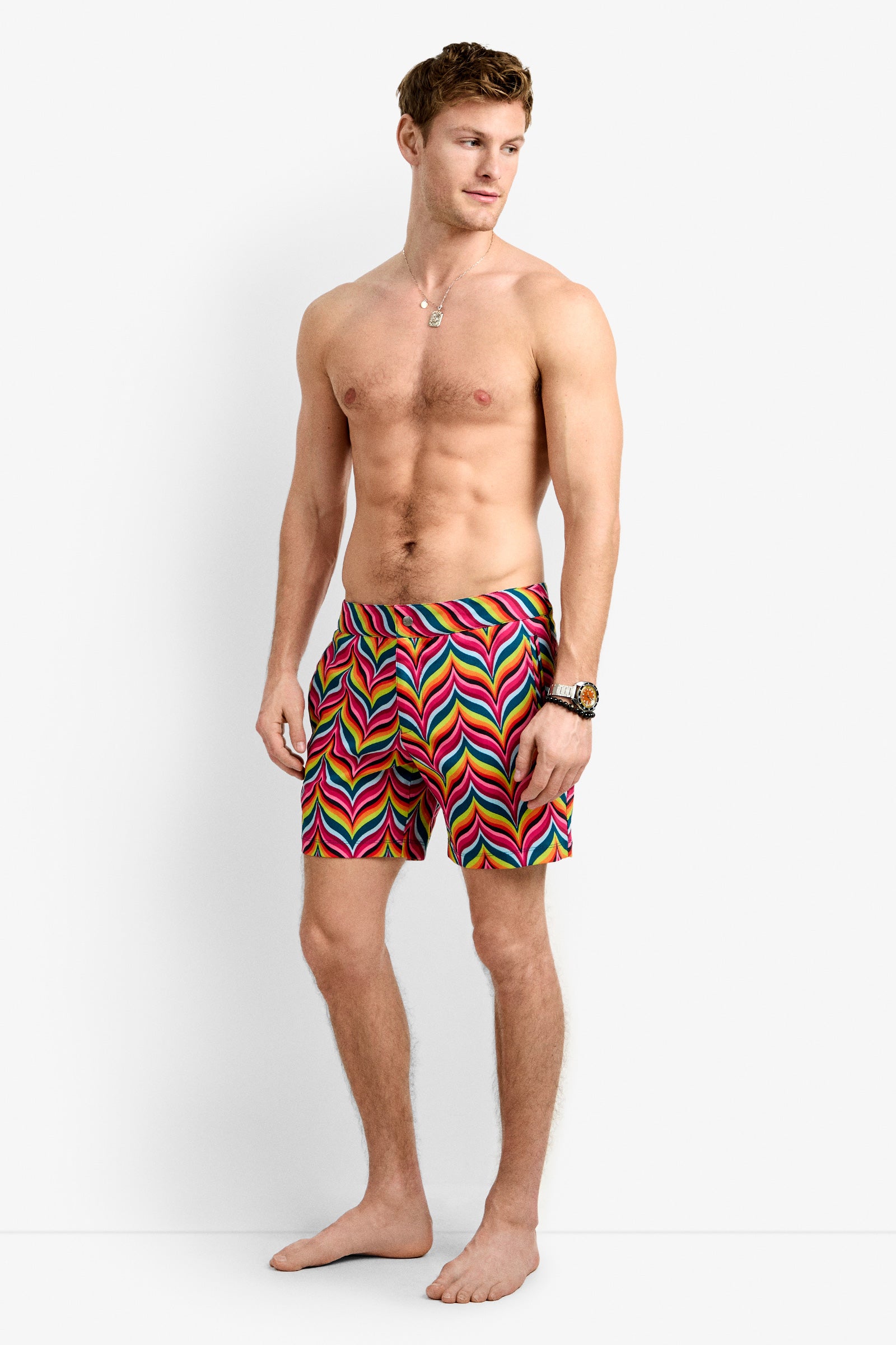 A shirtless man stands barefoot in the Honolulu Board Short - Chelsea Chevron, featuring a vibrant wavy multicolored design. He accessorizes with a necklace and watch, posing against a plain white background—fresh summer swim style.