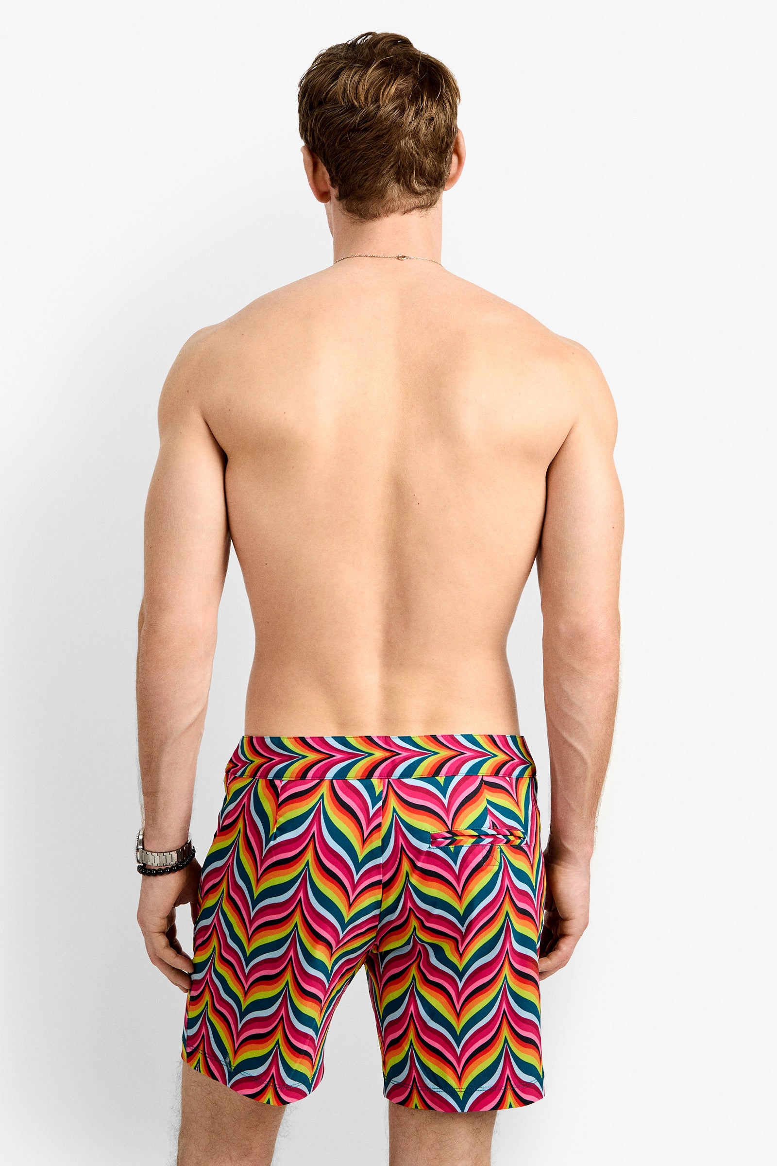 A shirtless man with short brown hair and a watch on his left wrist faces away, wearing Honolulu Board Short - Chelsea Chevron—vibrant swim trunks with a multicolor zigzag print, perfect for summer.