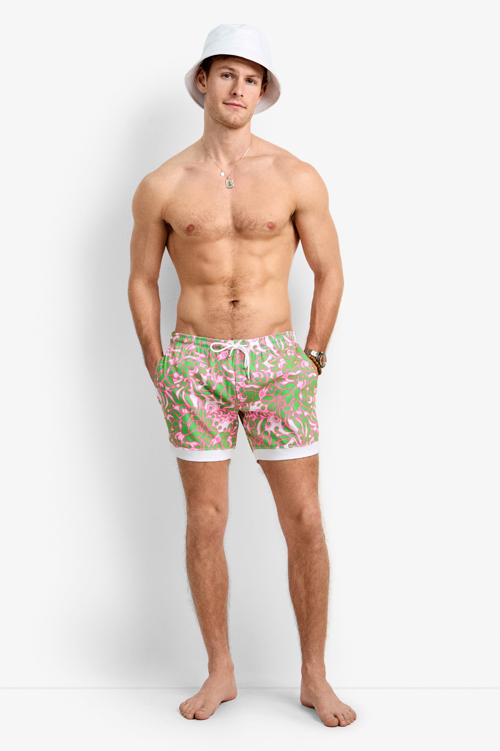 A shirtless man barefoot against a white background wears a white bucket hat, the Hermosa Beach Board Short - Petra Floral, a necklace, and a watch. His hands are in his pockets as he looks at the camera.