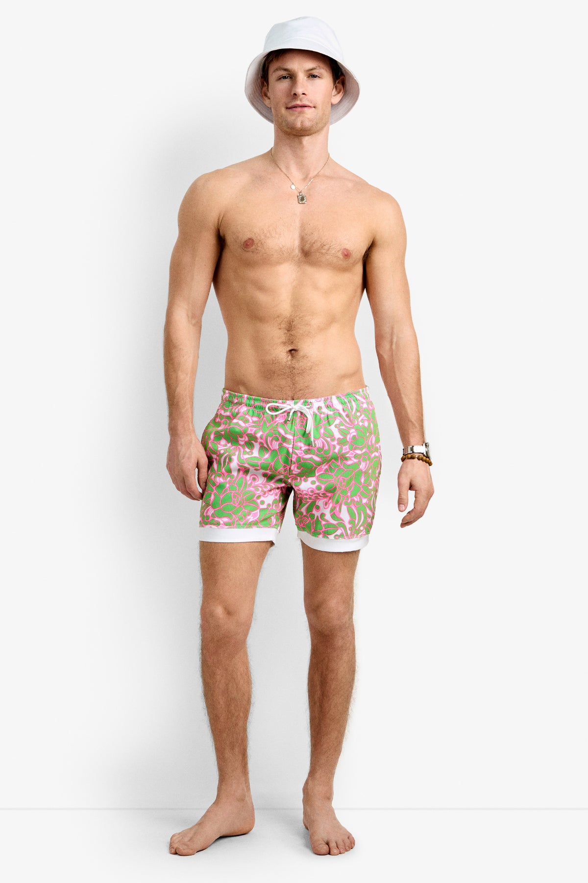 A shirtless man in a white bucket hat, gold necklace, watch, and Hermosa Beach Board Short - Petra Floral stands barefoot against a plain white background.