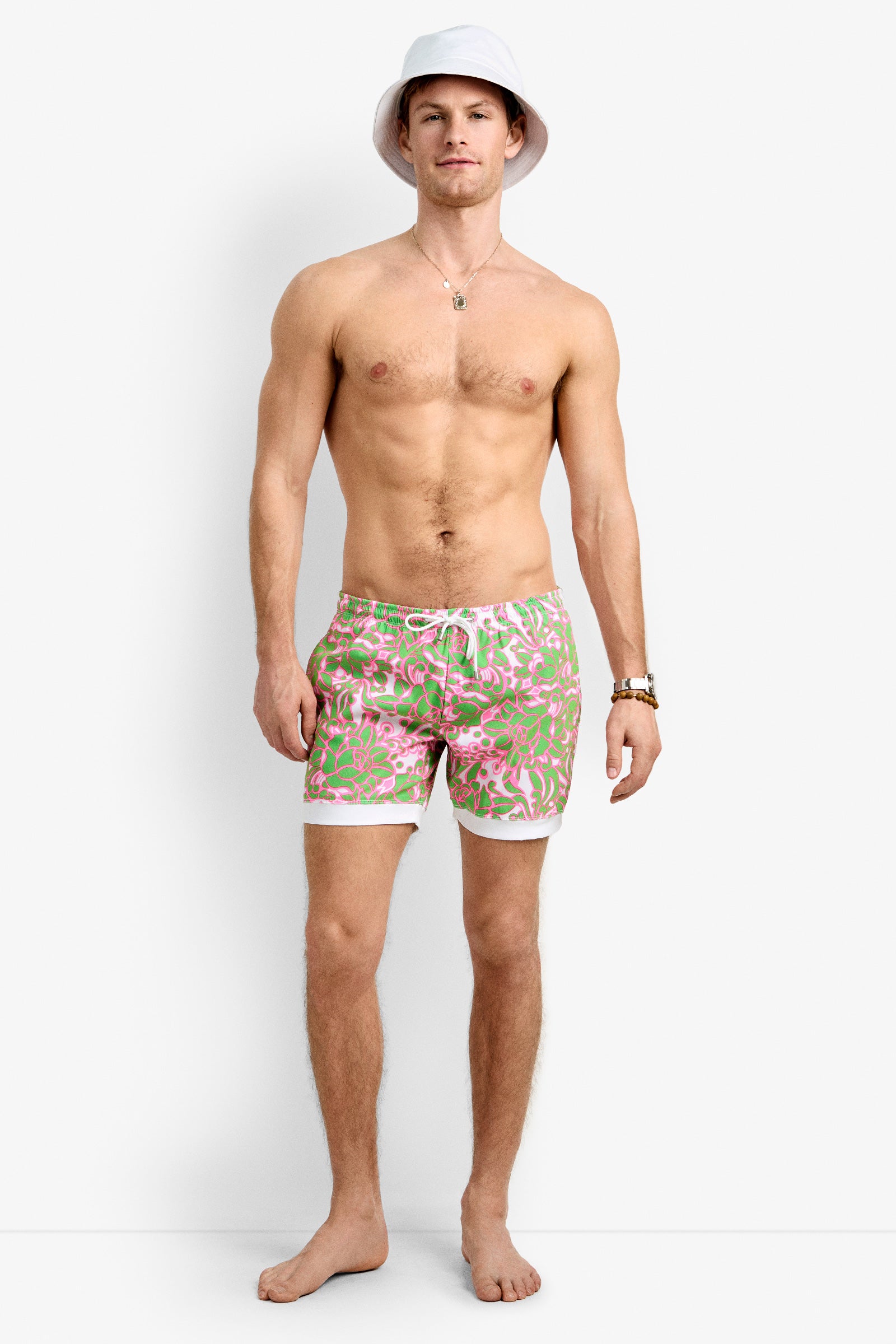 A shirtless man in a white bucket hat, gold necklace, watch, and Hermosa Beach Board Short - Petra Floral stands barefoot against a plain white background.