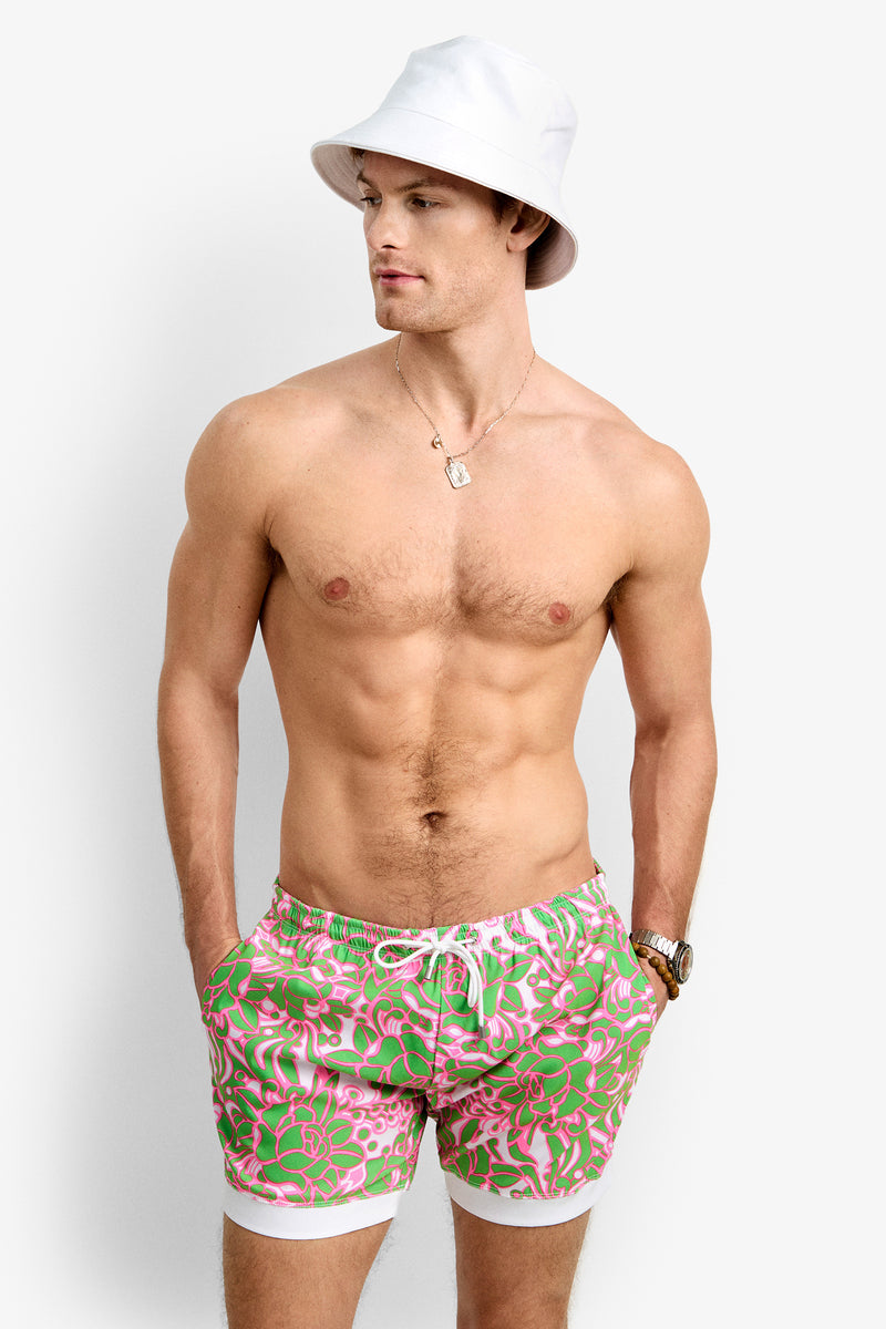 A shirtless man wears a white bucket hat, necklace, wristwatch, and Hermosa Beach Board Short - Petra Floral with green and pink floral patterns and white trim, standing with hands in pockets against a plain background.