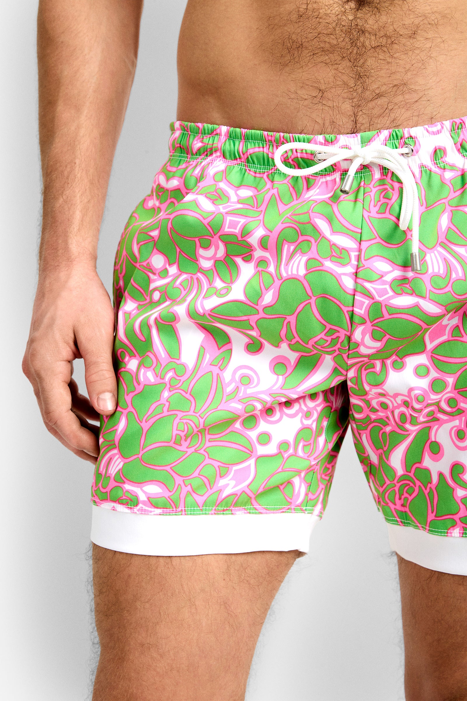 The lower torso and upper legs of a person wearing the Hermosa Beach Board Short - Petra Floral, featuring a bold green and pink abstract floral design with a white drawstring and white trim, are visible.