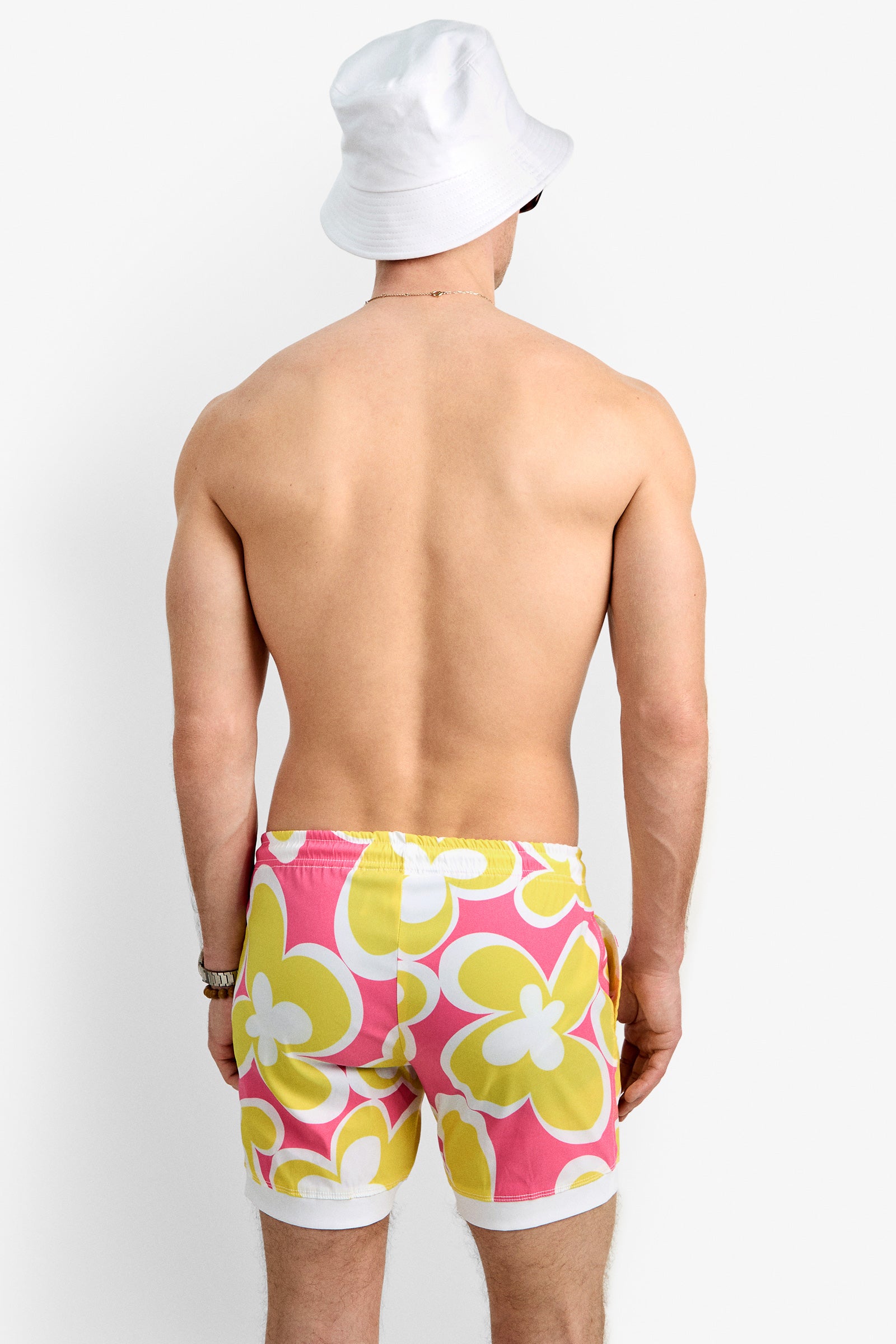 A man, shirtless and wearing a white bucket hat, models the Hermosa Beach Board Short—pink swim trunks with bold yellow and white flower patterns—against a plain white background.