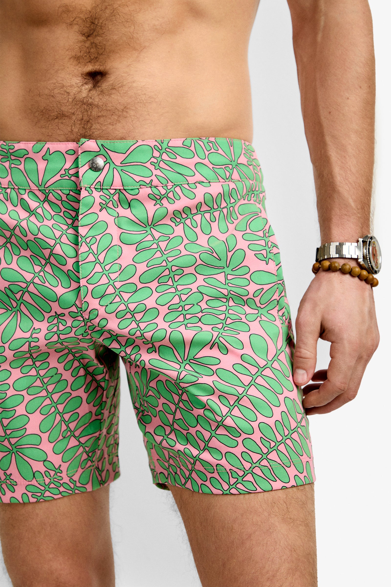 A man wears Honolulu Board Short - Vines, featuring a pink and green leaf pattern; only his torso and upper legs are shown, with a beaded bracelet and watch on his wrist.